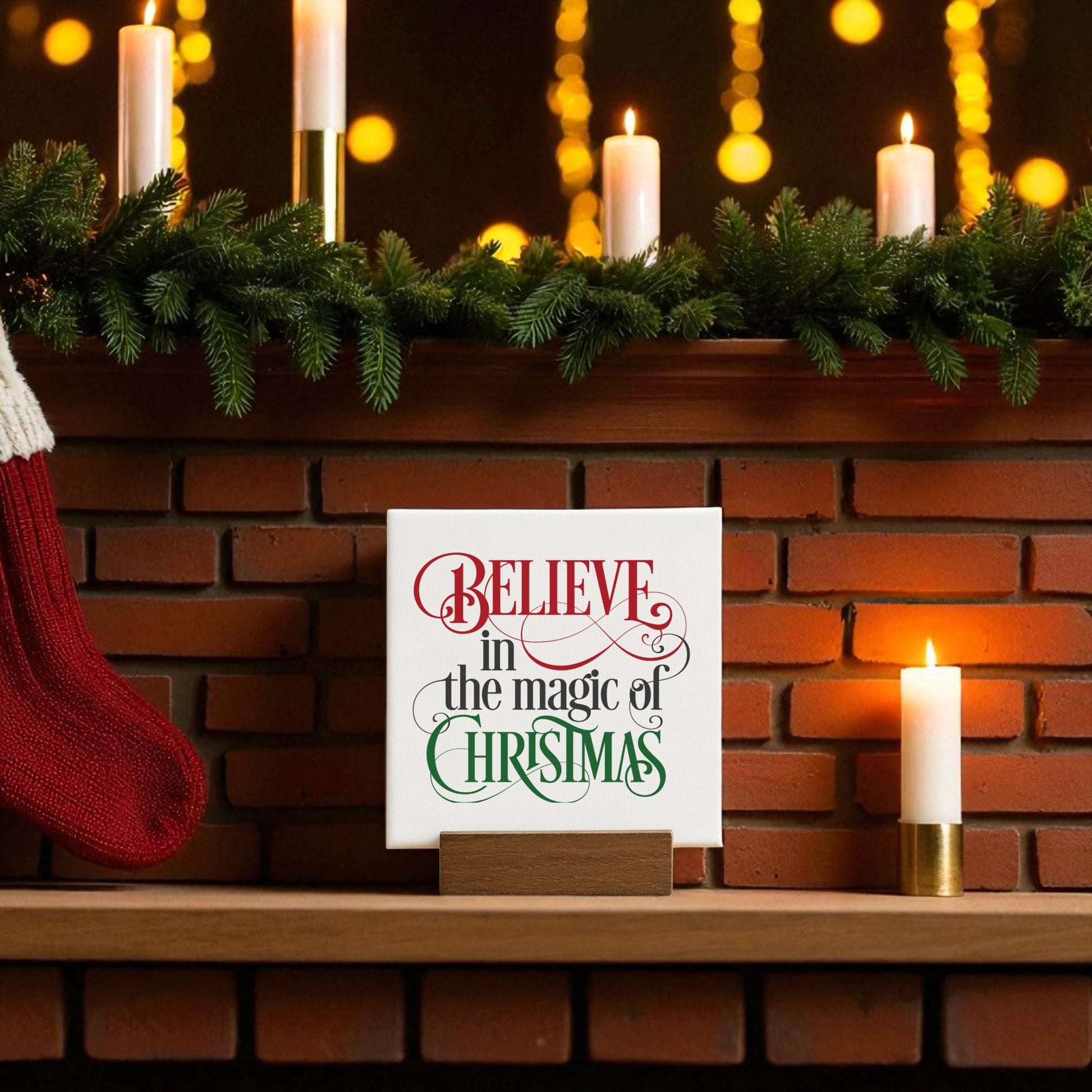 A festive mantel with evergreens, white candles, and a red stocking is complete with the Mirabilia Boutique "Believe in the Magic of Christmas" 4x4 mini sign, featuring red and green lettering for perfect holiday decor.