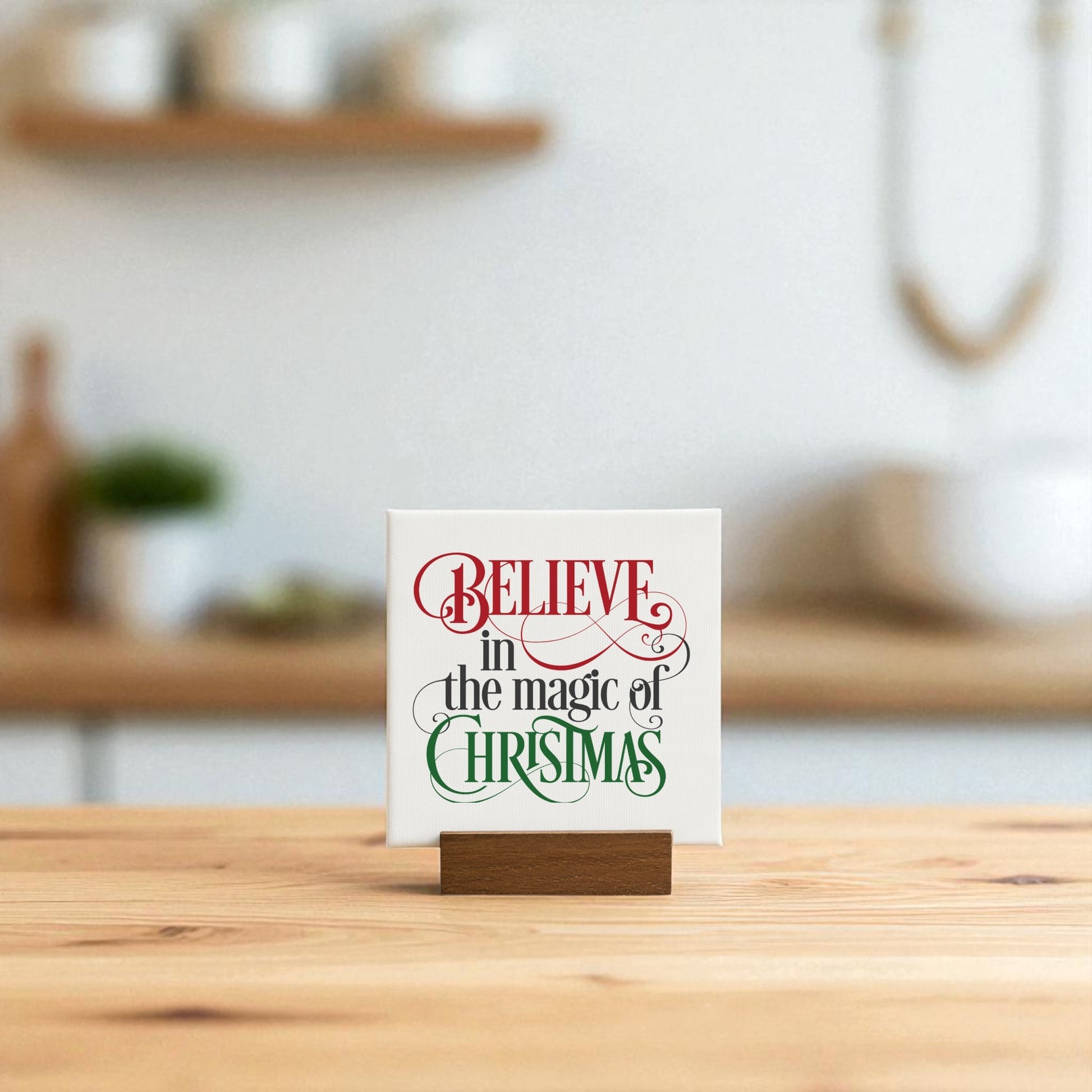 The Mirabilia Boutique "Believe in the Magic of Christmas" Mini Sign (4x4) sits on a table, displaying its festive red and green script. A softly blurred kitchen with holiday decor provides a cozy seasonal backdrop.