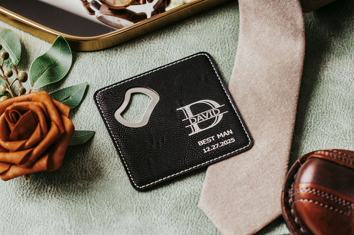 The Mirabilia Boutique Engraved Leatherette Bottle Opener Coaster makes a personalized best man gift, shown with “David,” “Best Man,” and “12-27-2025” engraving, resting on a green surface next to a brown rose, beige tie, and brown shoe.