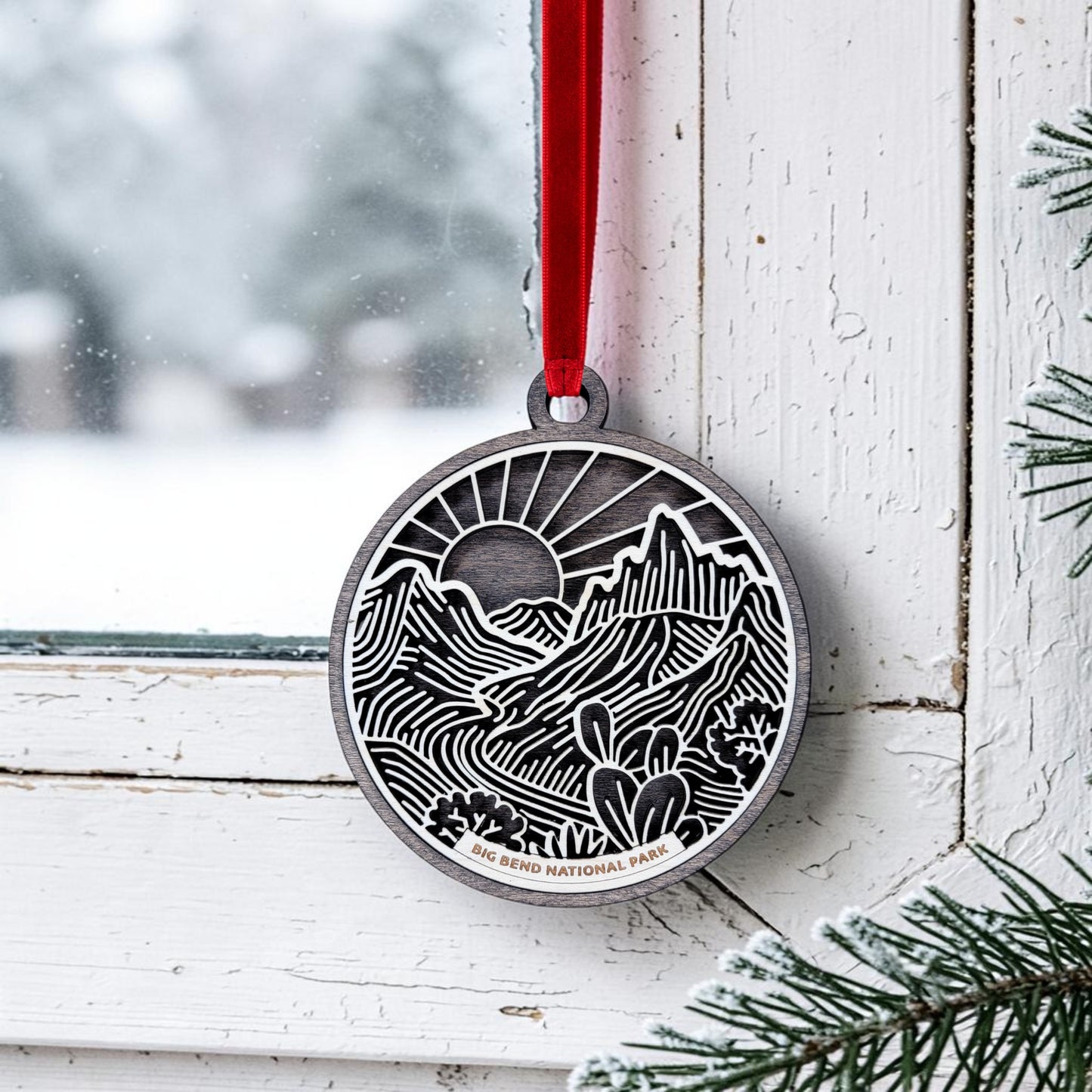 A Big Bend National Park Line Art Ornament by Mirabilia Boutique, showcasing intricate mountain, sun, and cactus designs in delicate Southwest line art, hangs from a red ribbon on a white window frame. Snow and pine branches are visible outside, while “Big Bend National Park” is engraved at the bottom of this unique metal keepsake.