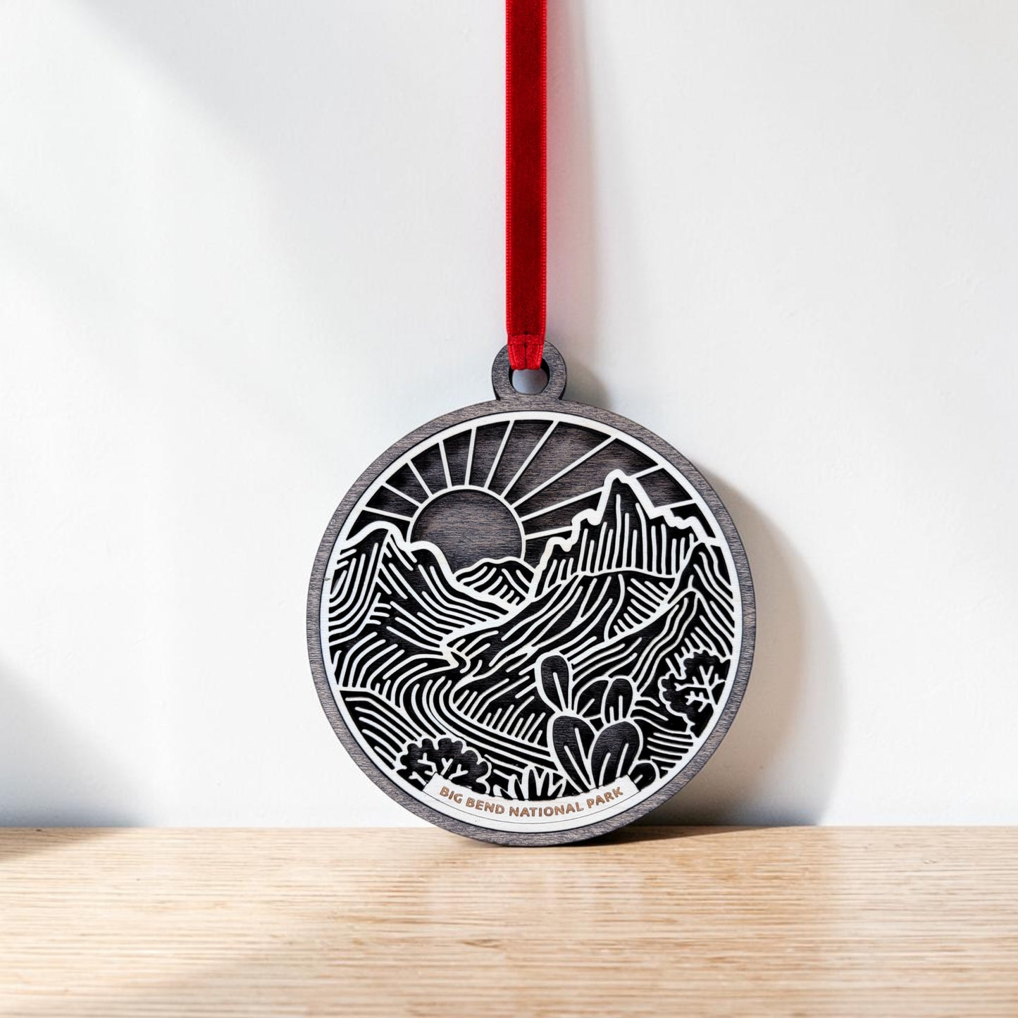 The Mirabilia Boutique Big Bend National Park Line Art Ornament—a round wooden keepsake adorned with intricate Southwest-inspired mountain, cactus, and sun designs—hangs from a red ribbon against a white background and rests on a light wooden surface.