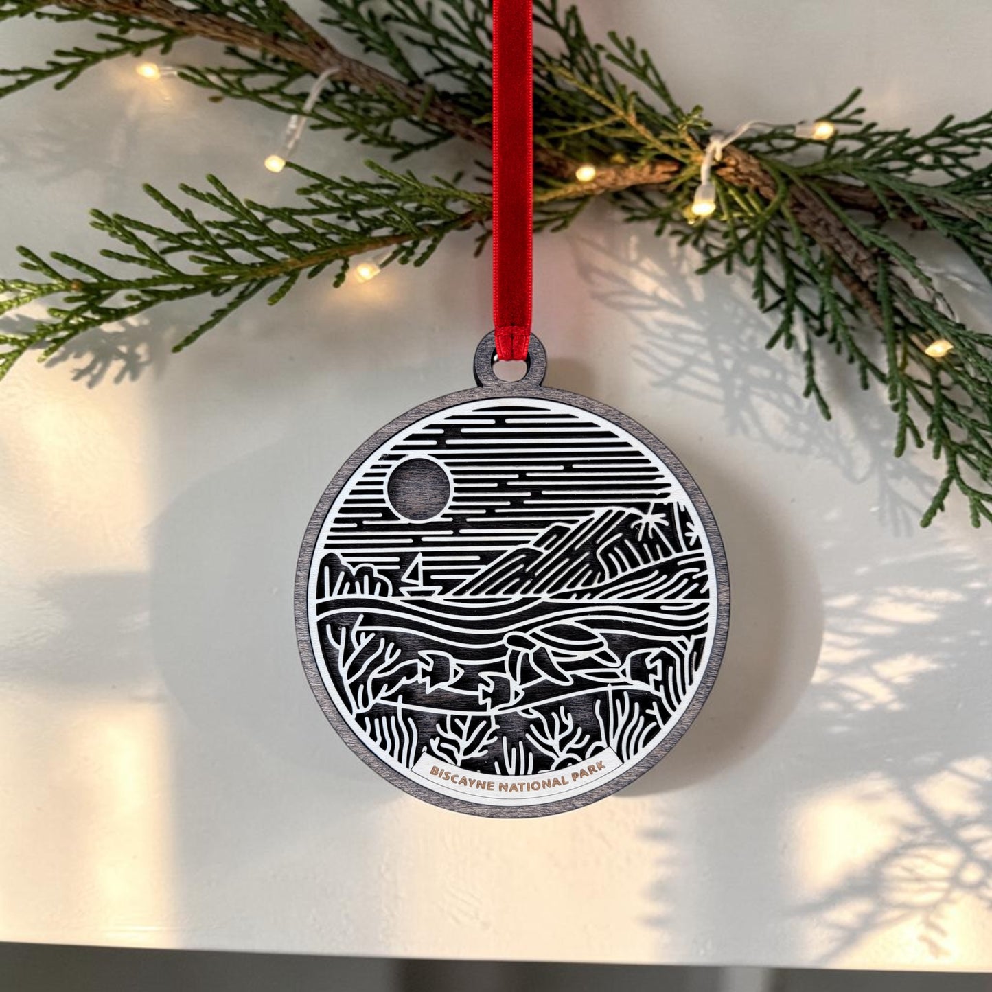 The Biscayne National Park Line Art Ornament by Mirabilia Boutique features a striking black and white line art design on a round wooden keepsake, elegantly hung from a red ribbon. Set amid pine branches decorated with delicate white lights, this unique ornament creates a festive and memorable display.