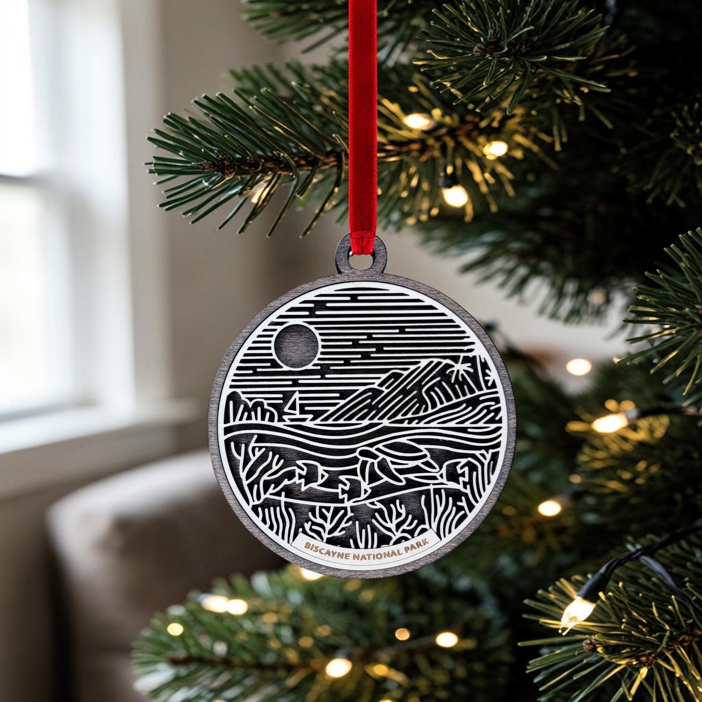 A round wooden Biscayne National Park Line Art Ornament by Mirabilia Boutique, featuring a black and white landscape design, hangs from a red ribbon on a lit Christmas tree. The ornament is inscribed with "Biscayne National Park" at the bottom, making it the perfect keepsake to celebrate your love for this stunning destination.