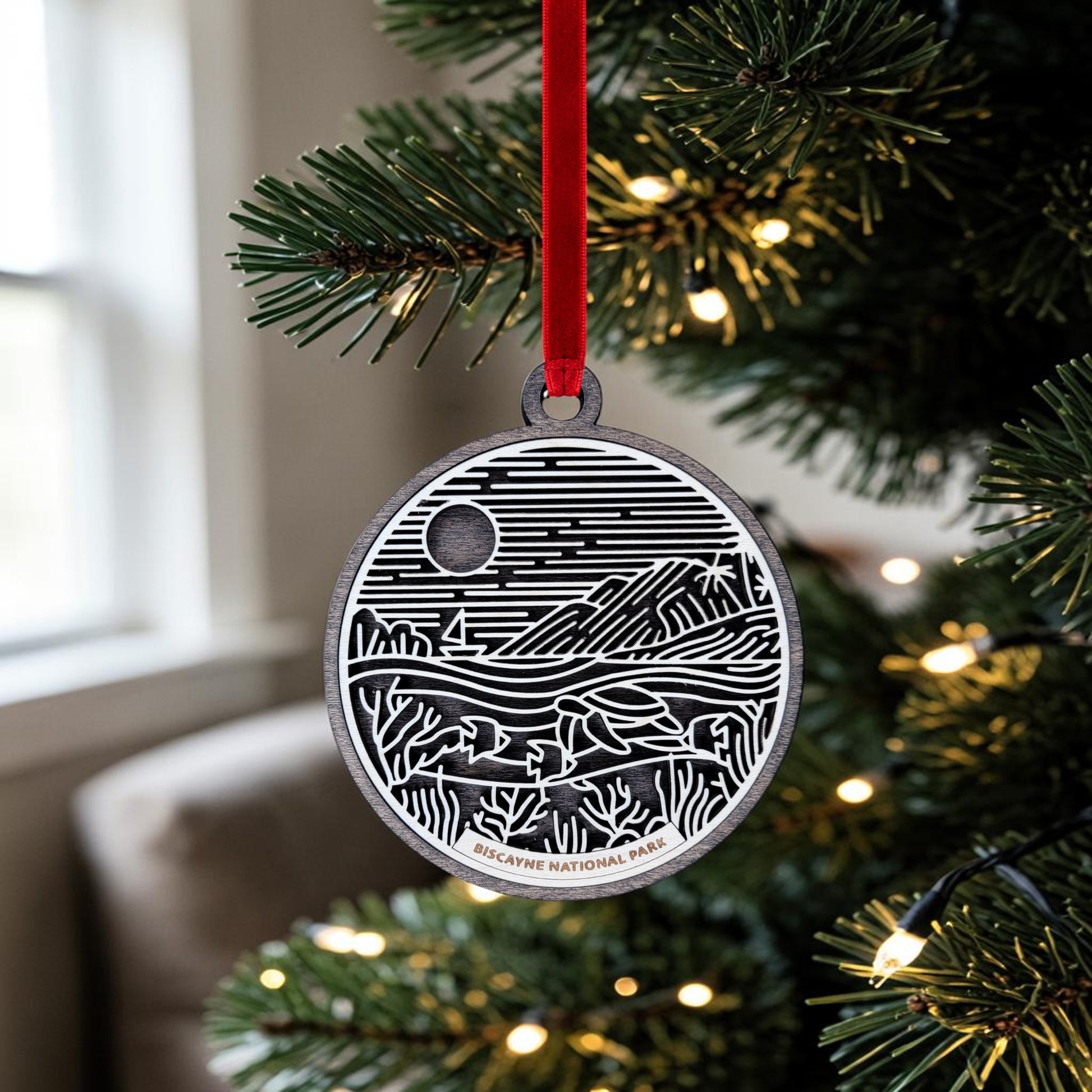 A round wooden Biscayne National Park Line Art Ornament by Mirabilia Boutique, featuring a black and white landscape design, hangs from a red ribbon on a lit Christmas tree. The ornament is inscribed with "Biscayne National Park" at the bottom, making it the perfect keepsake to celebrate your love for this stunning destination.