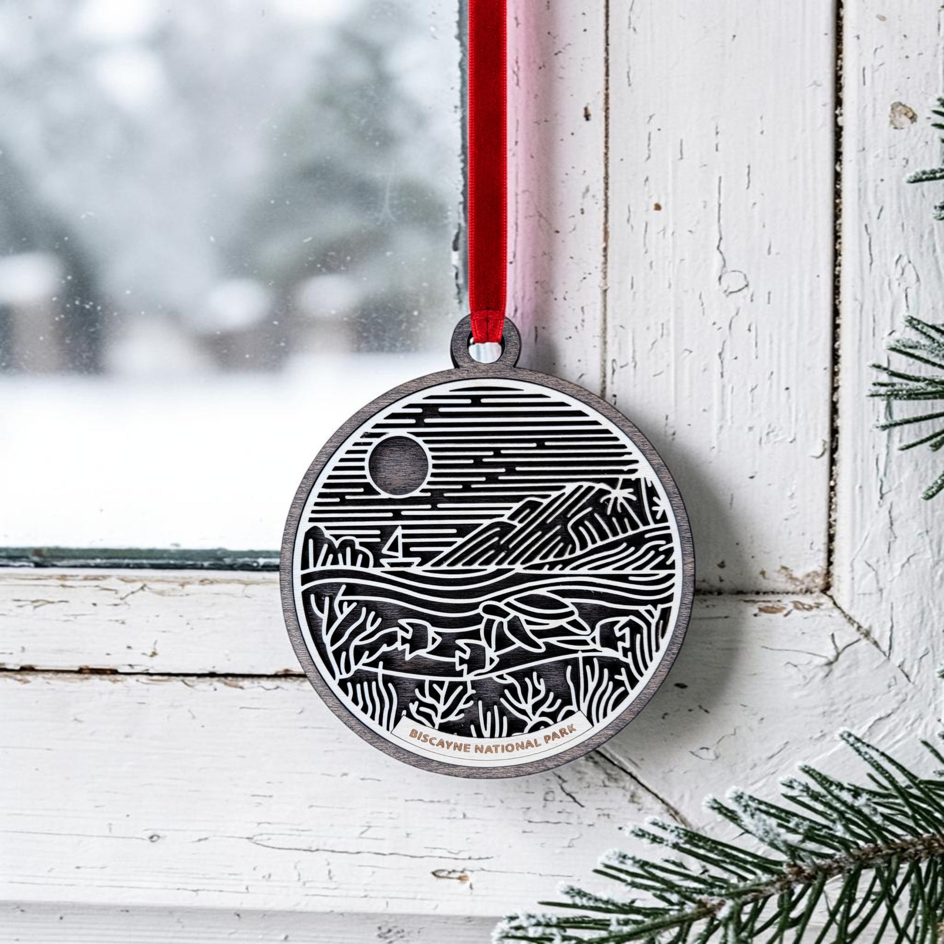 A round wooden Biscayne National Park Line Art Ornament by Mirabilia Boutique, featuring a black and white landscape design, is hung by a red ribbon on a window latch. Snowy scenery can be seen outside, with pine branches in the corner, and "Biscayne National Park" is inscribed on the ornament.