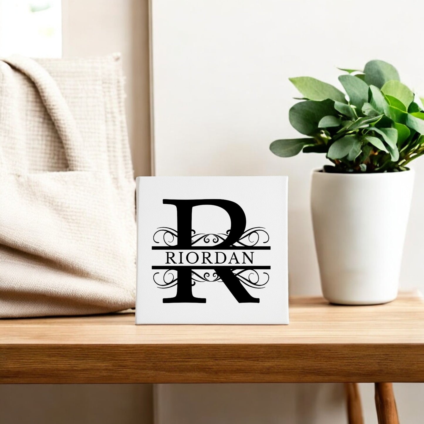 The Mirabilia Boutique Custom Monogram Canvas with Family Name – 8x8 features a bold black "R" and "RIORDAN" in decorative text, displayed on a white square canvas next to a beige bag and a potted green plant.