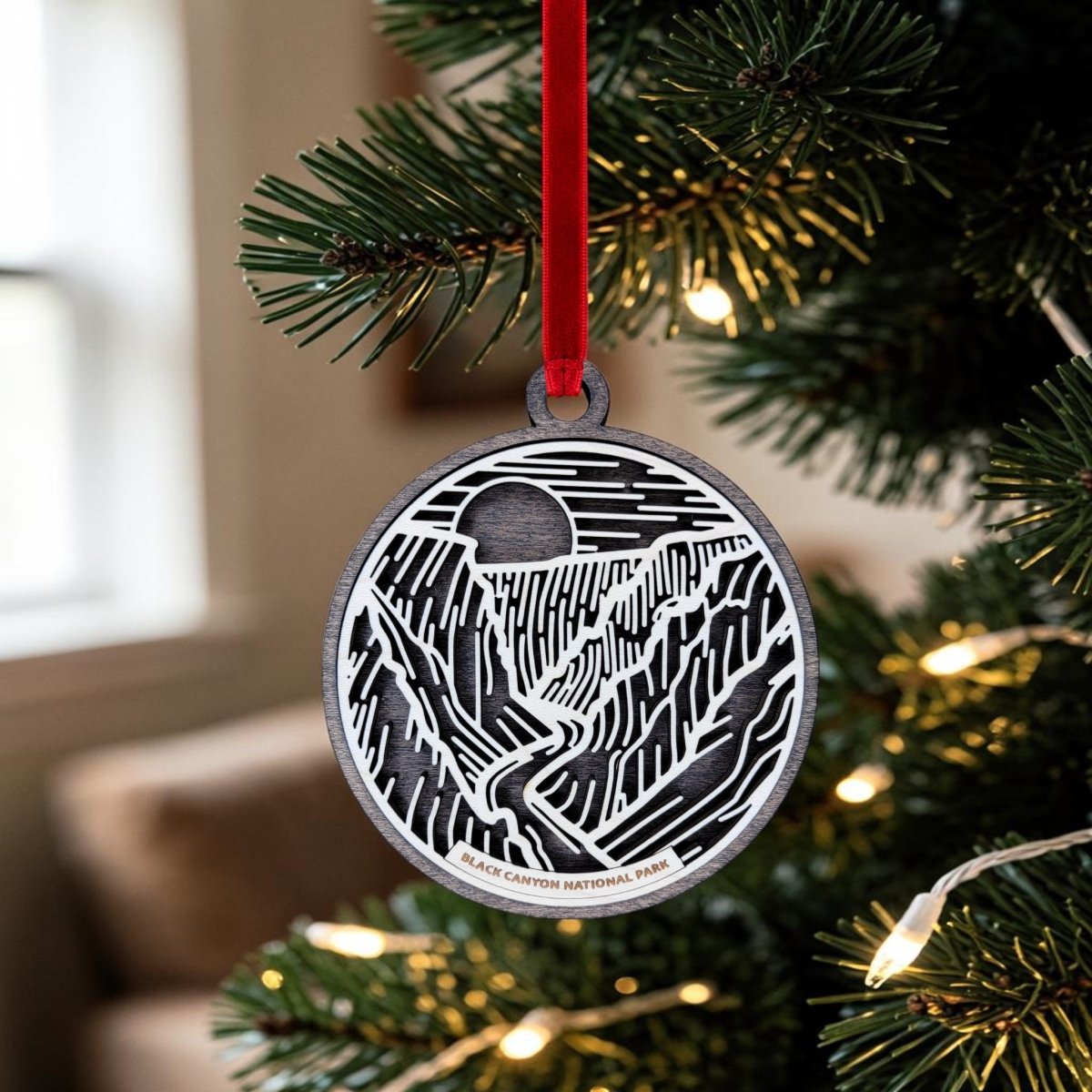 A Black Canyon of The Gunnison National Park Christmas Ornament by Mirabilia Boutique, featuring a black and white engraved mountain landscape, hangs from a red ribbon on a Christmas tree adorned with green needles and white lights.