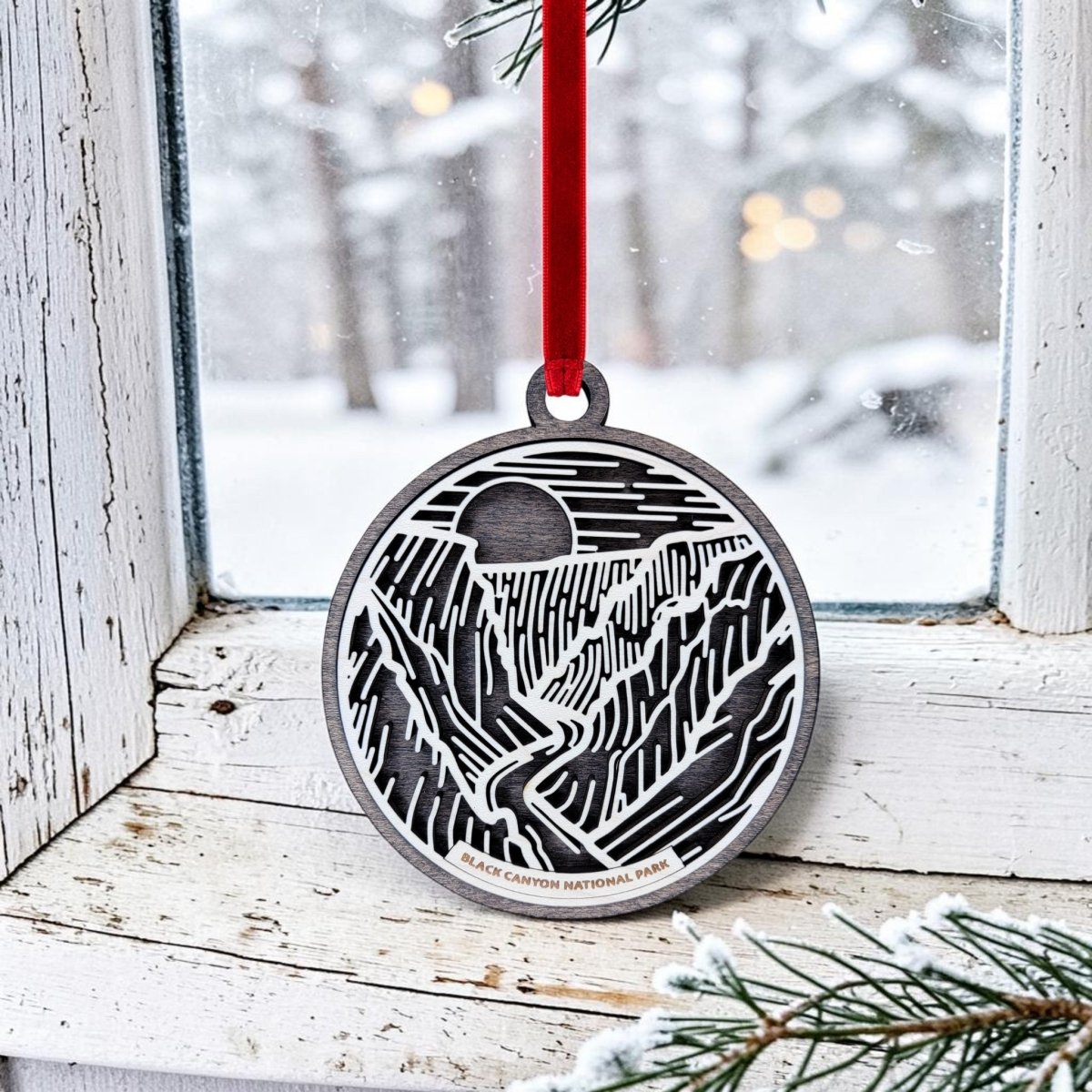 A Mirabilia Boutique "Black Canyon of The Gunnison National Park Christmas Ornament"—handcrafted from wood and adorned with black and white line art of the park—hangs by a red ribbon on a frosty window, with snow-covered trees and gentle lights shining outside.