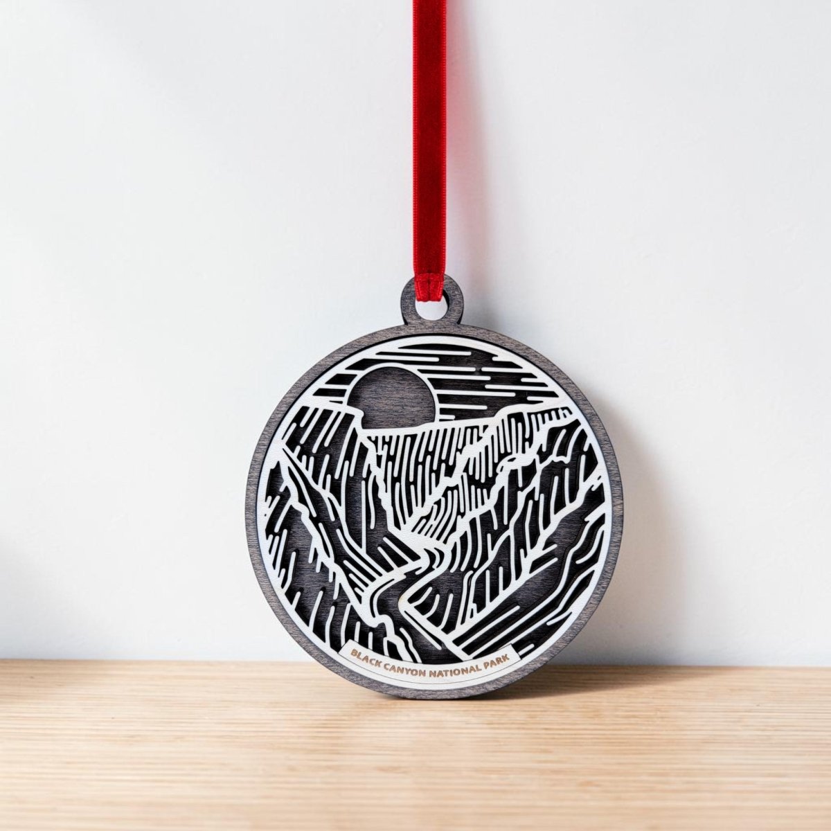 Introducing the Black Canyon of The Gunnison National Park Christmas Ornament by Mirabilia Boutique—a handcrafted wooden ornament featuring a delicately carved canyon landscape with a river and sun or moon motif. Suspended by a red ribbon, the base is inscribed with "Black Canyon of the Gunnison National Park." This distinctive line art ornament from Mirabilia Boutique is showcased against a crisp white background.