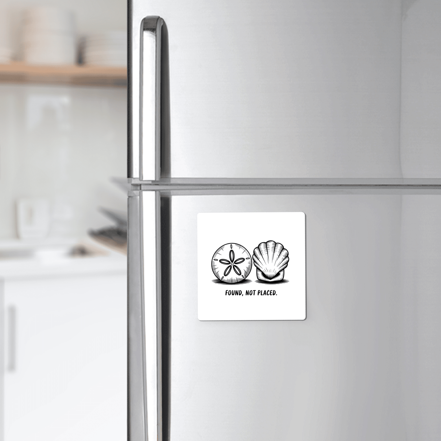 A stainless steel fridge displays the “Found, Not Placed Magnet – Minimalist Coastal Quote” by Mirabilia Boutique, featuring a hand-drawn shell and sand dollar. The kitchen with beach decor and open shelves appears in the background.