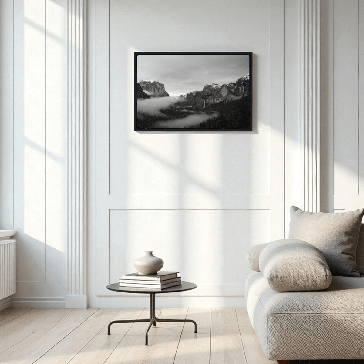 A minimal living room features a light sofa, round black coffee table, and the Yosemite Valley Black & White Fine Art Framed Print by Mirabilia Boutique above. White paneled walls and a large window fill the space with natural light.