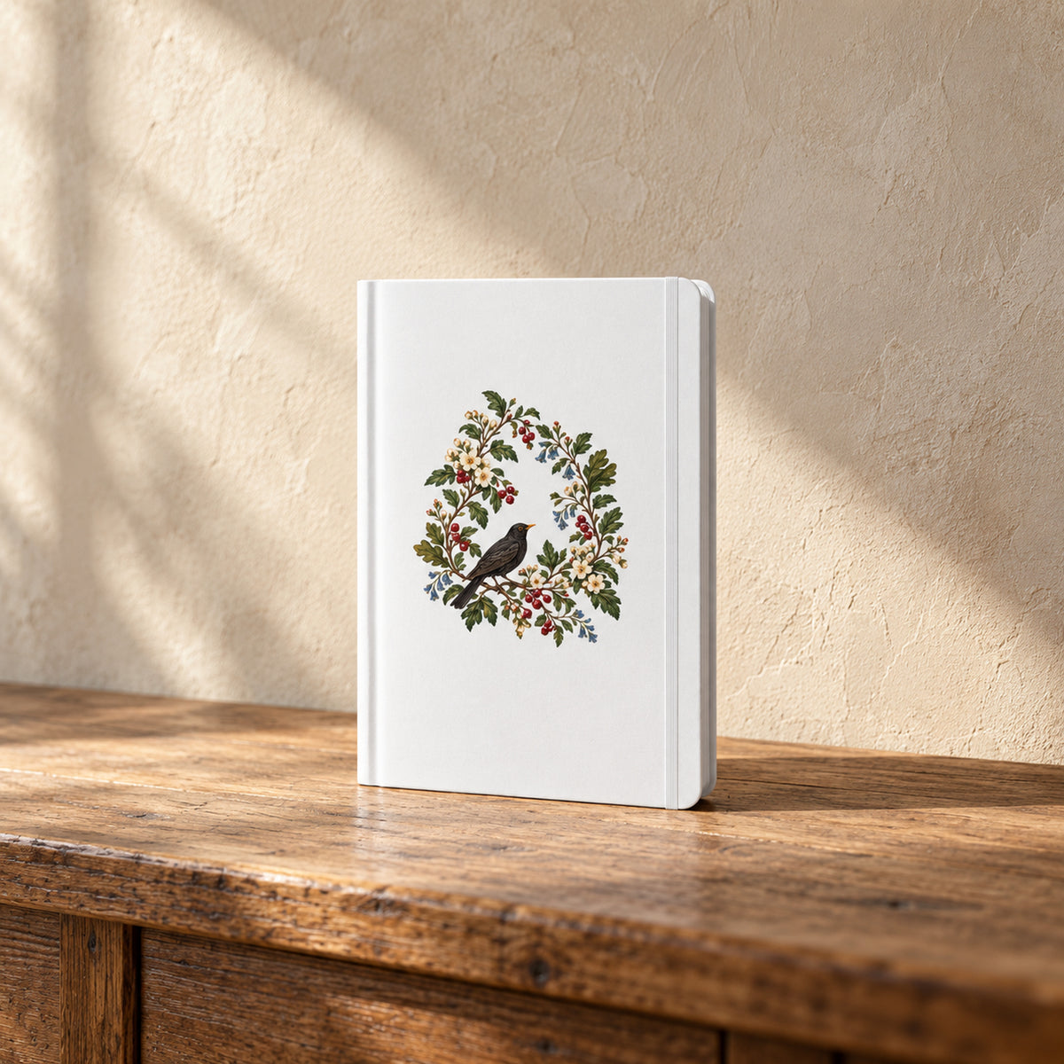 The Garden Ledger A5 Lined Journal by Mirabilia Boutique stands upright, featuring a central illustration of a black bird perched in a botanical wreath of berries and flowers on a white cover.