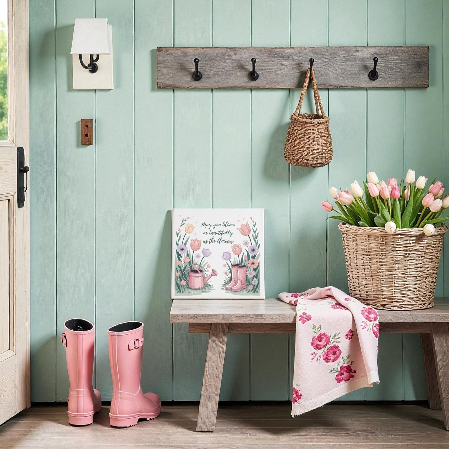 A cozy entryway with mint green walls, a wooden bench, pink rain boots, pink tulips in a basket, and spring decor—a Mirabilia Boutique "Bloom Like the Flowers Canvas Art – Spring Garden Quote" hanging above in a wicker bag on wall hooks.