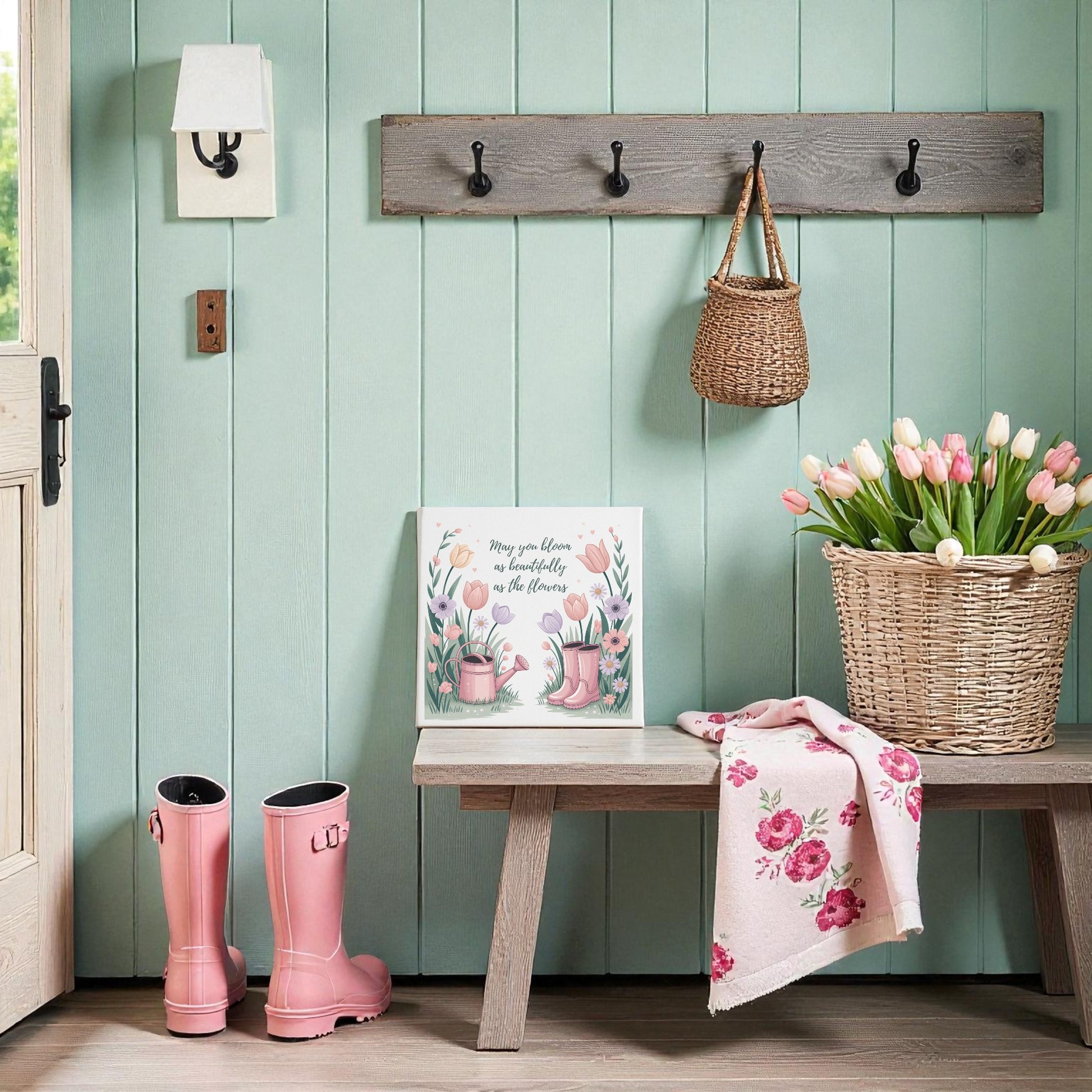 A cozy entryway with mint green walls, a wooden bench, pink rain boots, pink tulips in a basket, and spring decor—a Mirabilia Boutique "Bloom Like the Flowers Canvas Art – Spring Garden Quote" hanging above in a wicker bag on wall hooks.
