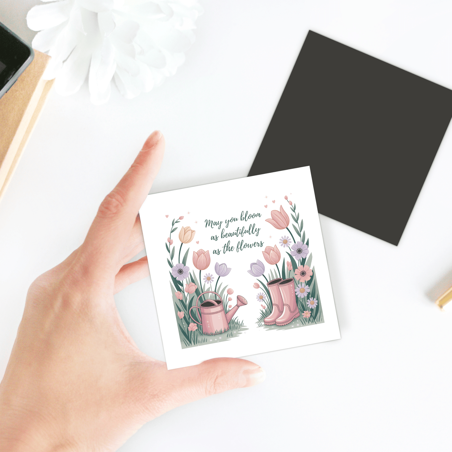 A hand holds the Mirabilia Boutique "Bloom Like the Flowers Magnet – Spring Floral Quote," featuring boots, flowers, and a watering can illustration with the quote, “May you bloom as beautifully as the flowers.” A pen and white flower are nearby.