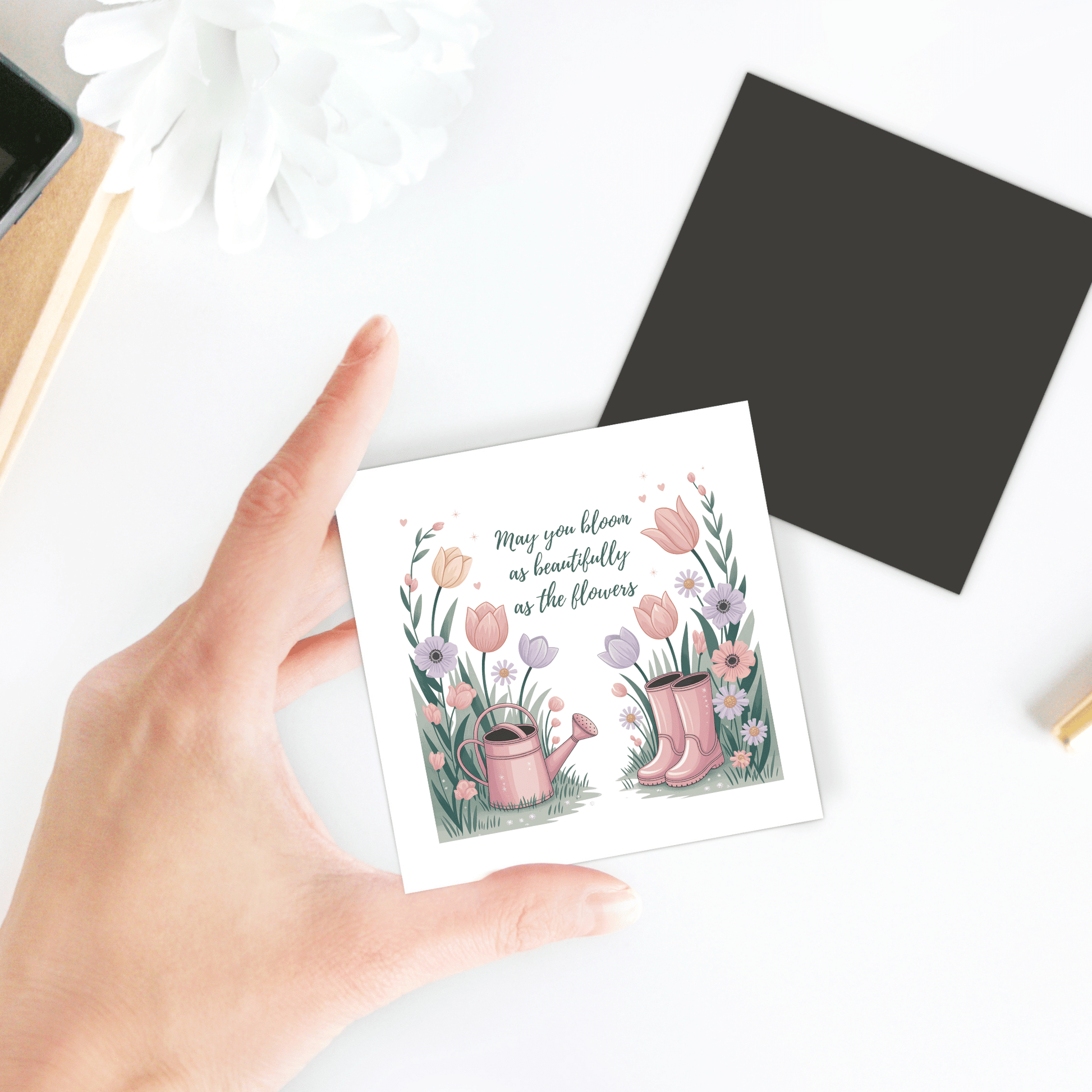 A hand holds the Mirabilia Boutique "Bloom Like the Flowers Magnet – Spring Floral Quote," featuring boots, flowers, and a watering can illustration with the quote, “May you bloom as beautifully as the flowers.” A pen and white flower are nearby.