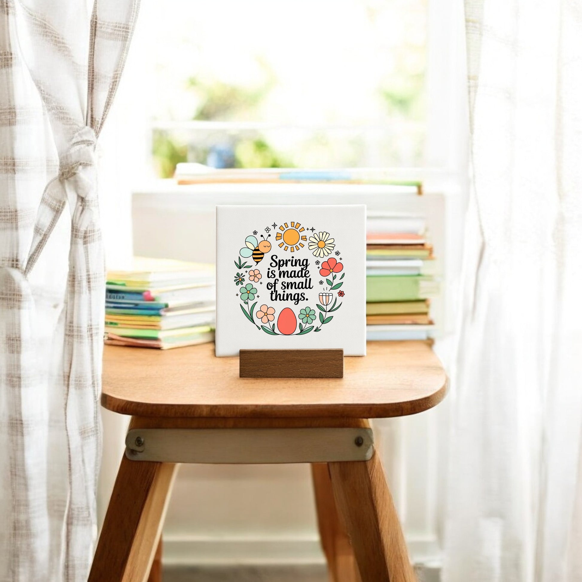 The Mirabilia Boutique "Spring Is Made of Small Things" Mini Canvas Sign sits on a wooden stool, featuring colorful hand-drawn floral wreaths. Stacks of books and sheer curtains complete the cozy spring decor scene.