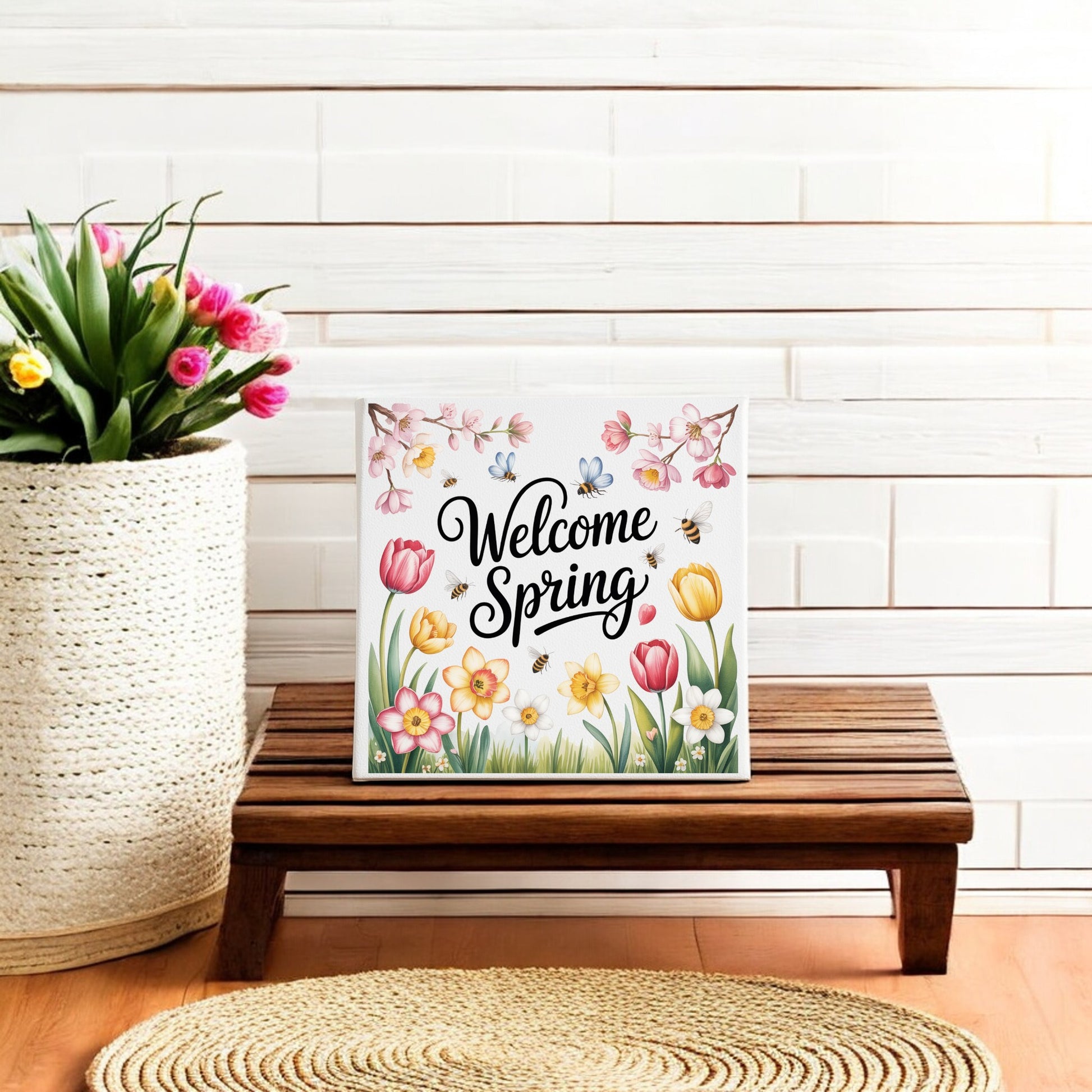 The Mirabilia Boutique Welcome Spring Canvas – 8x8 Floral Wall Art features flowers and butterflies, creating charming seasonal decor on a white shiplap wall beside a basket of pink and yellow tulips.
