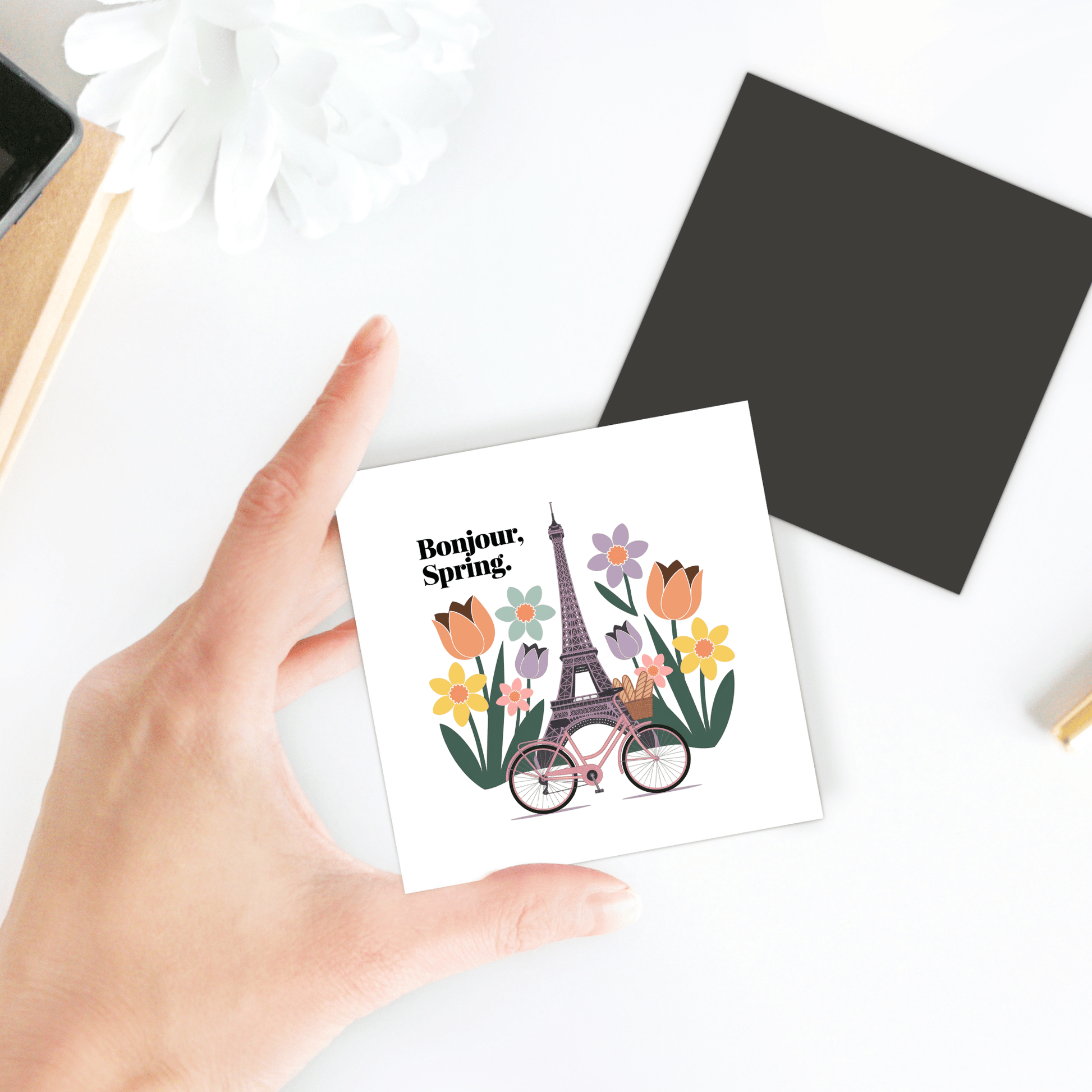 A hand holds the Bonjour Spring Magnet – Paris Bicycle & Floral Charm by Mirabilia Boutique, featuring a bike, flowers, and Eiffel Tower design—perfect for Parisian kitchen decor. In the background: a white flower, black square, and part of a desk.