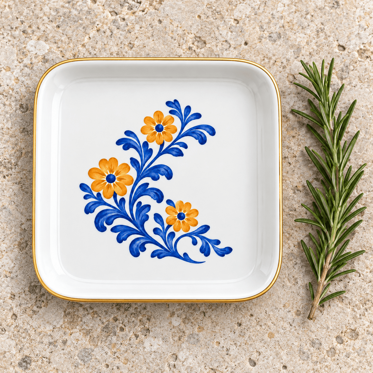 Courtyard Bloom Jewelry Tray, a square ceramic dish with a gold rim and painted blue and orange floral design, next to a sprig of rosemary.