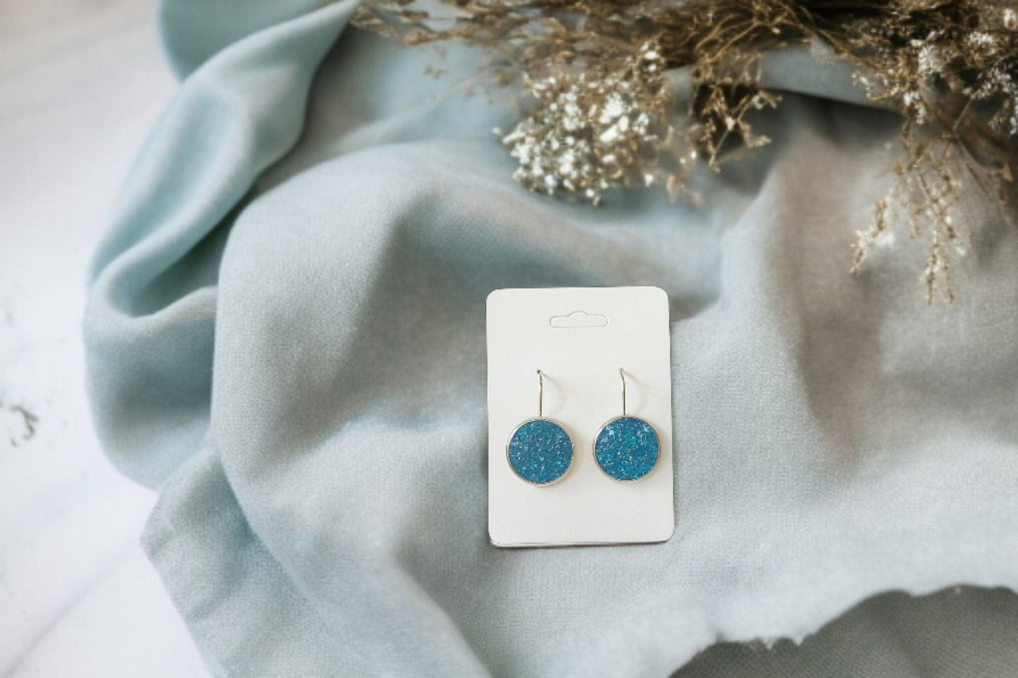 Blue Crushed Glass Earrings by Mirabilia Boutique rest on a white card atop light blue fabric with dried flowers—these silver dangle earrings are perfect for women who love unique jewelry.