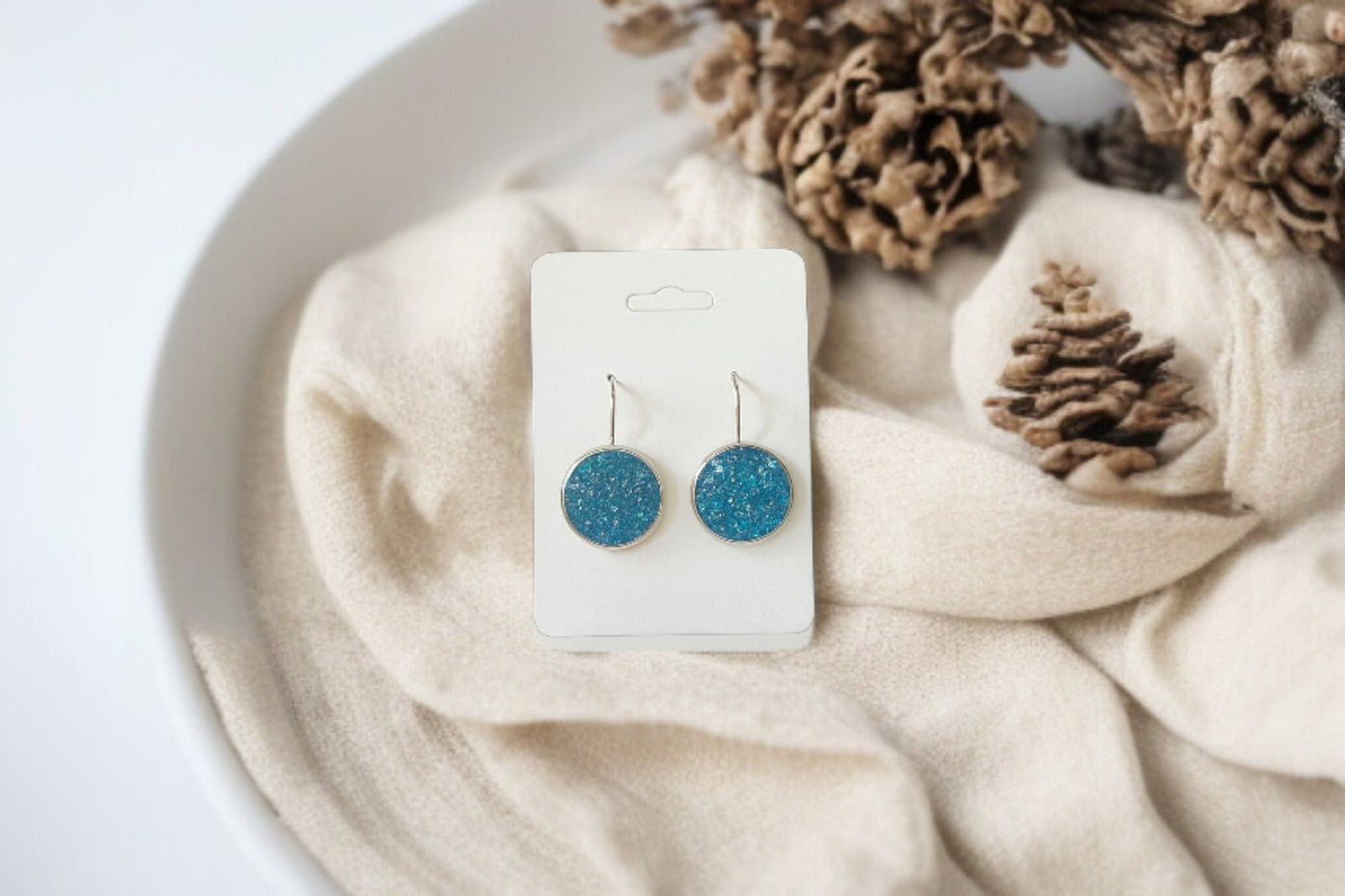 Mirabilia Boutique’s Blue Crushed Glass Earrings | Silver Dangle Earrings for Women are shown on a white card atop beige fabric in a white bowl with pinecones—perfect for anyone seeking unique jewelry.