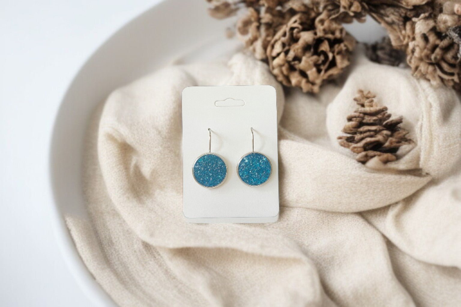 Mirabilia Boutique’s Blue Crushed Glass Earrings | Silver Dangle Earrings for Women are shown on a white card atop beige fabric in a white bowl with pinecones—perfect for anyone seeking unique jewelry.