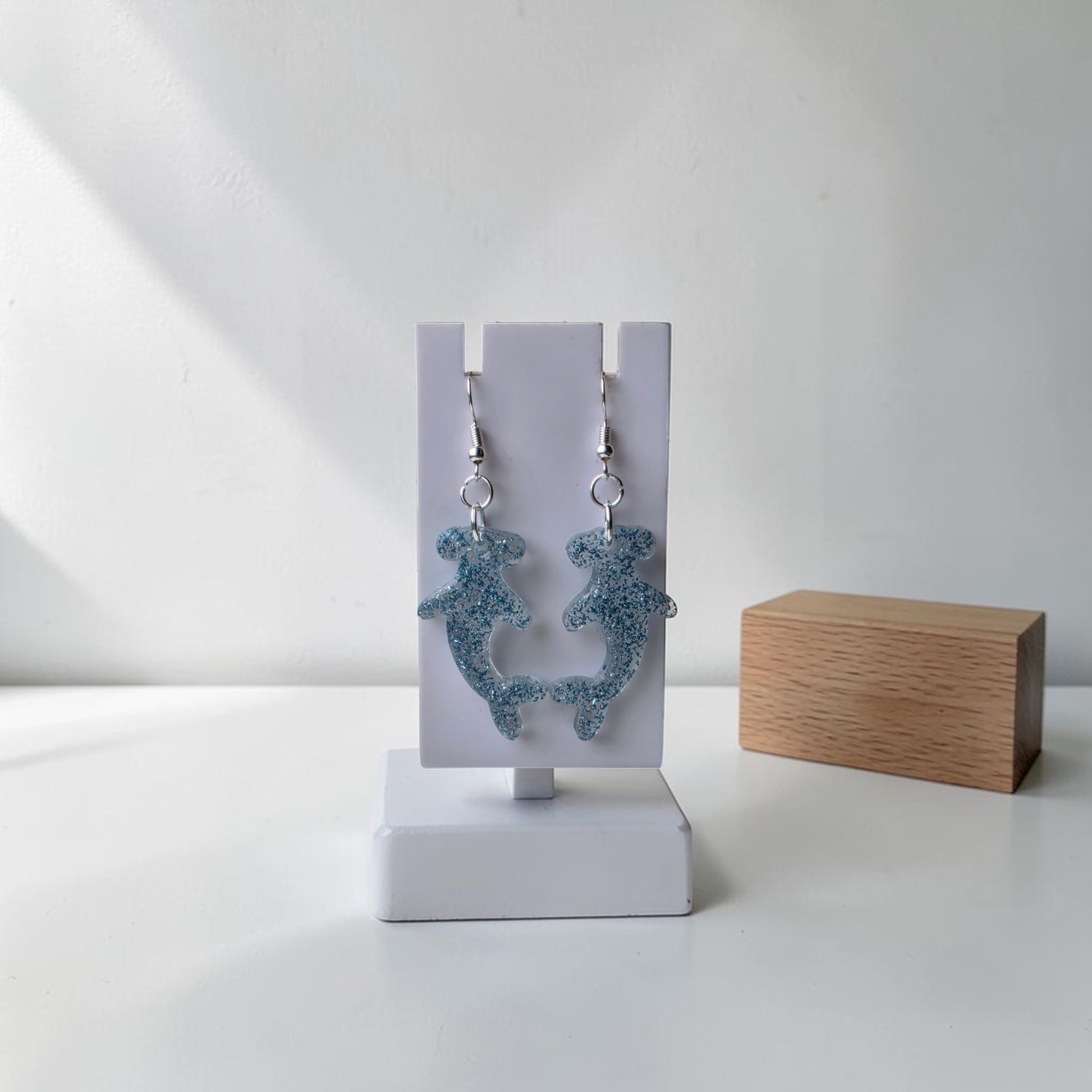 Mirabilia Boutique’s Blue Glitter Hammerhead Shark Earrings hang on a white display stand with a wooden block in the background—ideal ocean-inspired jewelry to enhance any collection.