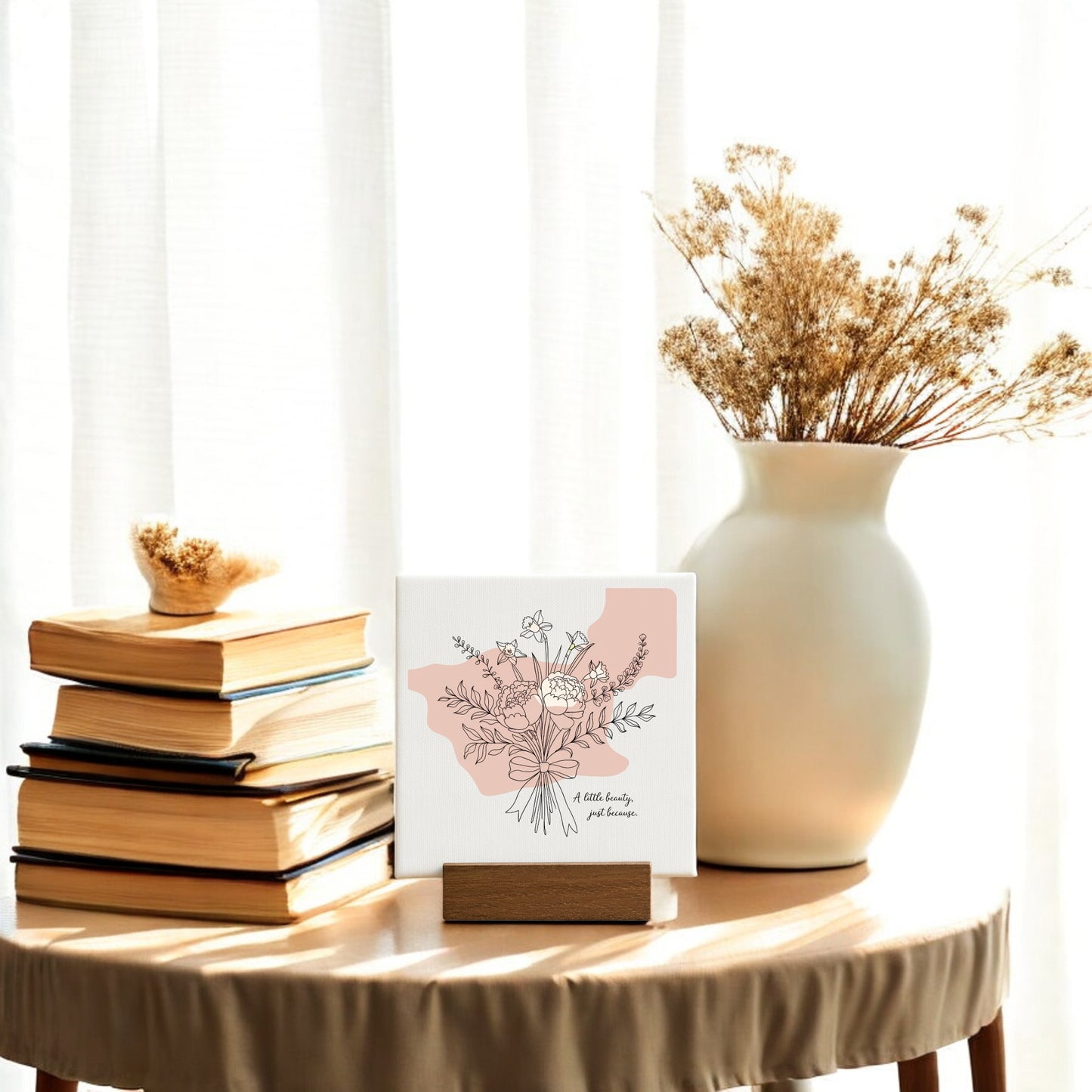 A small table with a stack of books, a white vase with dried flowers, and the Mirabilia Boutique "A Little Beauty Mini Canvas – Just Because Floral Sign" sits by a window—perfect for minimalist decor or as a thoughtful gift.