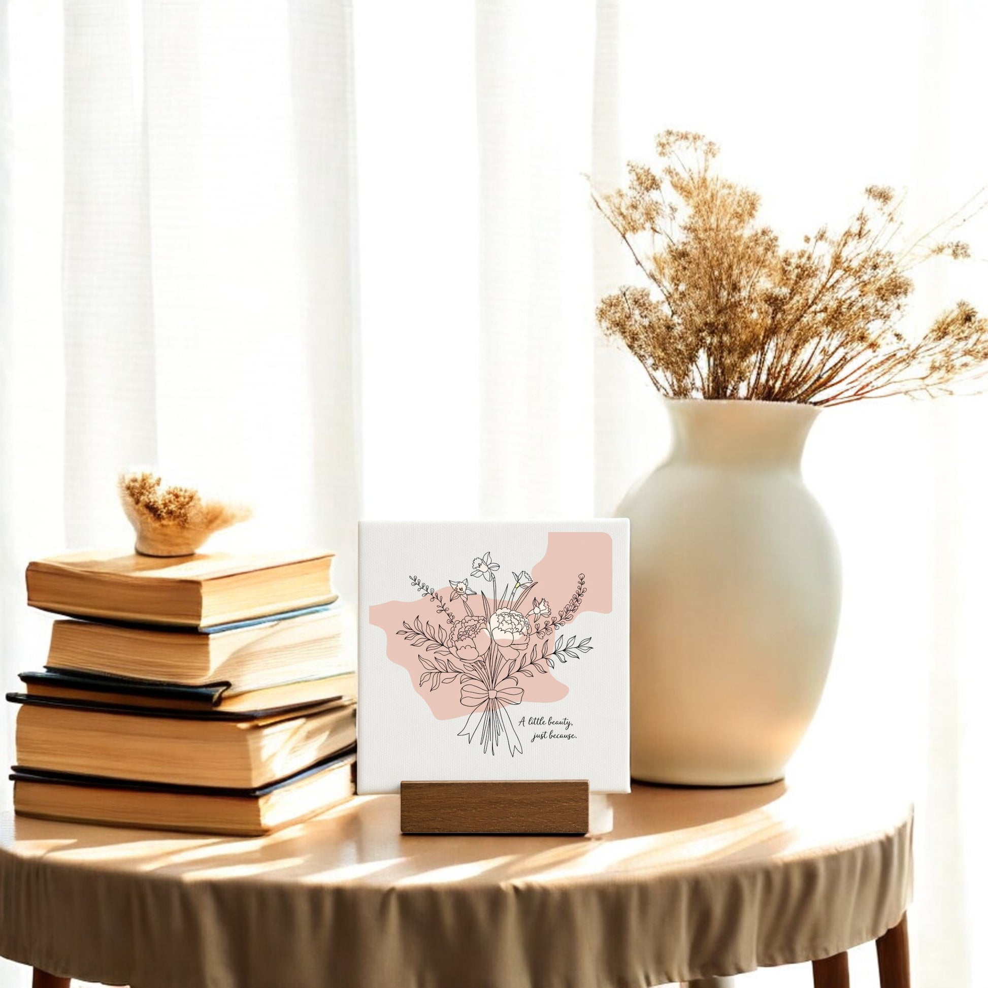 A small table with a stack of books, a white vase with dried flowers, and the Mirabilia Boutique "A Little Beauty Mini Canvas – Just Because Floral Sign" sits by a window—perfect for minimalist decor or as a thoughtful gift.