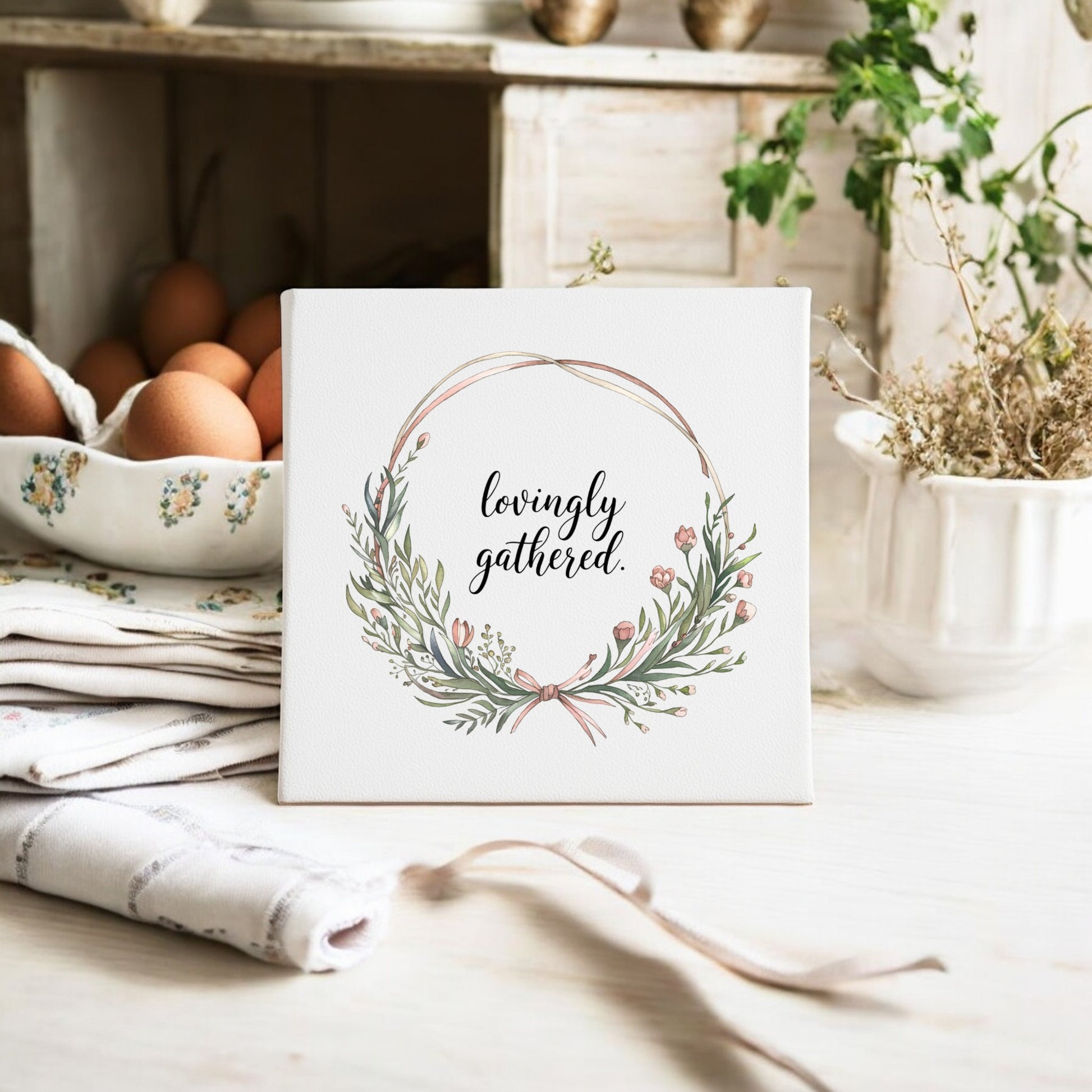 The Mirabilia Boutique Lovingly Gathered Canvas – 8x8 Botanical Wreath Wall Art, featuring a floral wreath and “lovingly gathered,” is displayed on a kitchen counter beside eggs, folded linens, and dried herbs.