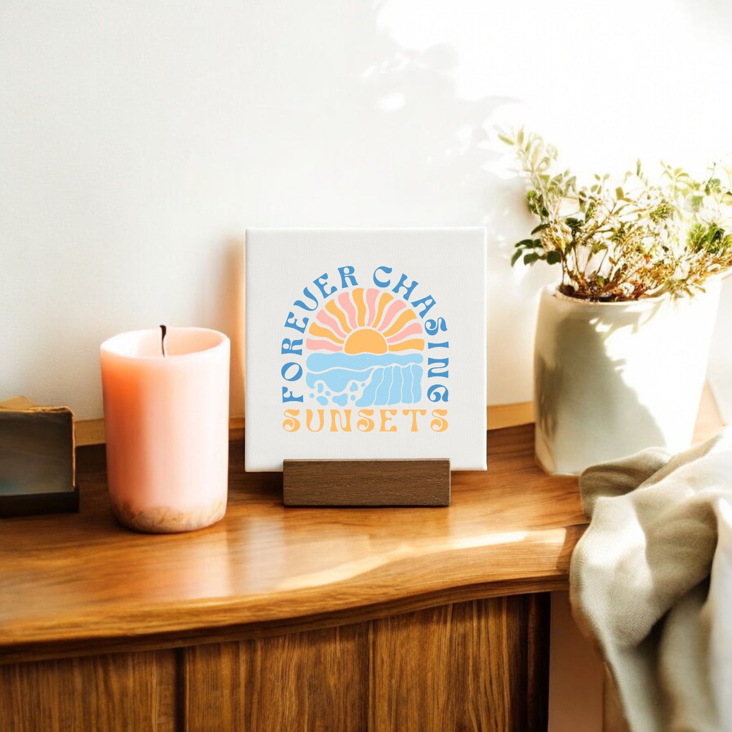 A wooden table displays a pink candle, potted plant, and Mirabilia Boutique's "Forever Chasing Sunsets Mini – Boho Pastel Canvas" with sunset wall art, all lit warmly by sunlight in this charming decor scene.