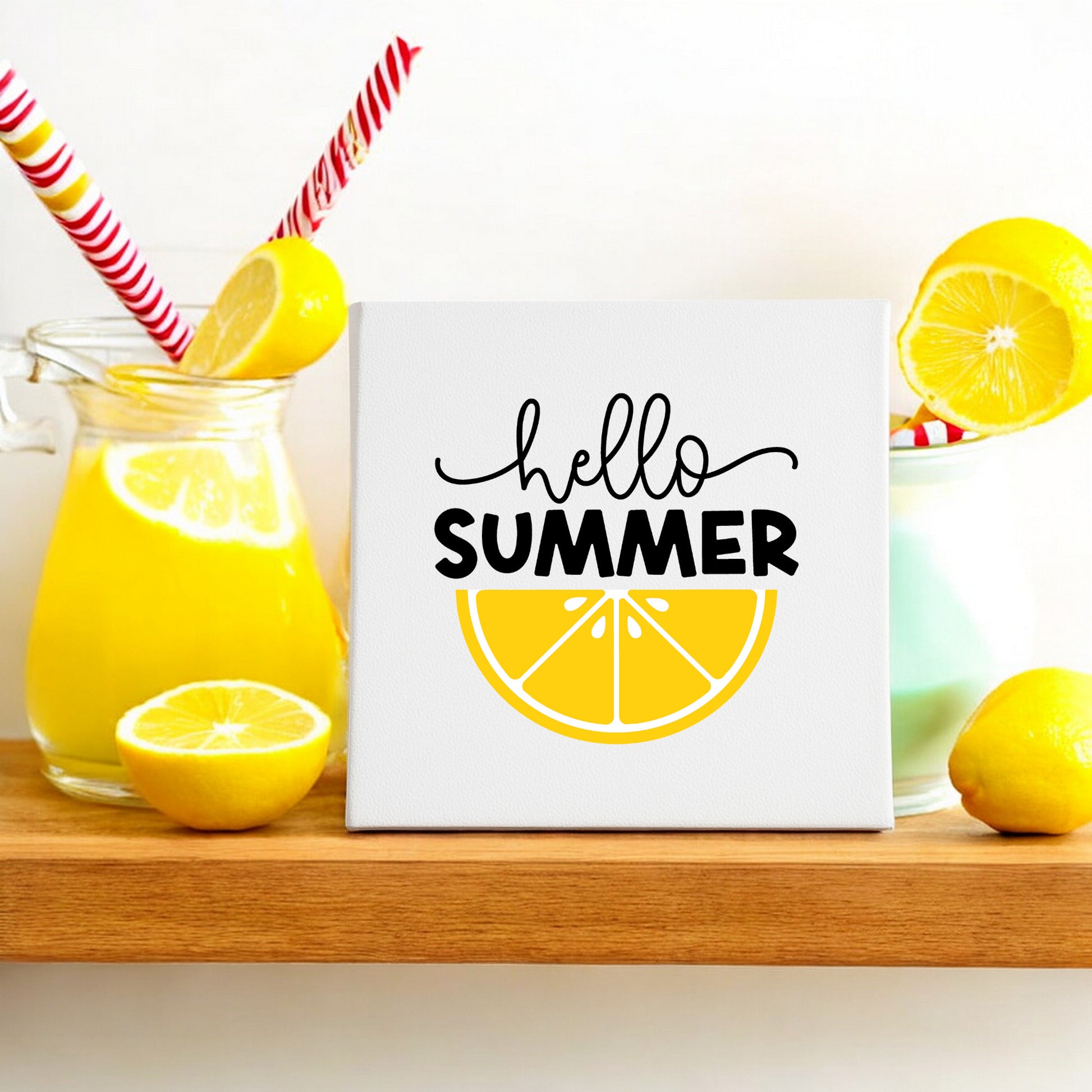 A jug and glass of yellow lemonade with striped straws sit by lemons on a wooden shelf. The Mirabilia Boutique Hello Summer Lemon Canvas Sign (8x8 Wall Art) with lemon slice art adds a cheerful touch to the décor.