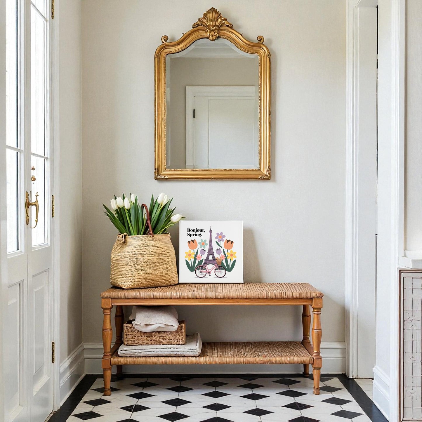 A hallway features a woven bench with a basket of white tulips, stacked towels, and Mirabilia Boutique's Bonjour, Spring Canvas – Eiffel Tower Floral Art. Above the bench hangs a gold ornate mirror; the floor displays black and white checkered tile.