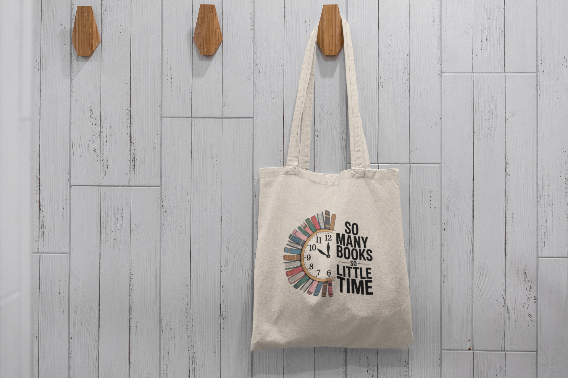 The So Many Books Canvas Tote Bag by Mirabilia Boutique, featuring a clock-shaped stack of books and "So many books, so little time," hangs on a wooden hook—perfect as a book lovers gift or stylish library tote.