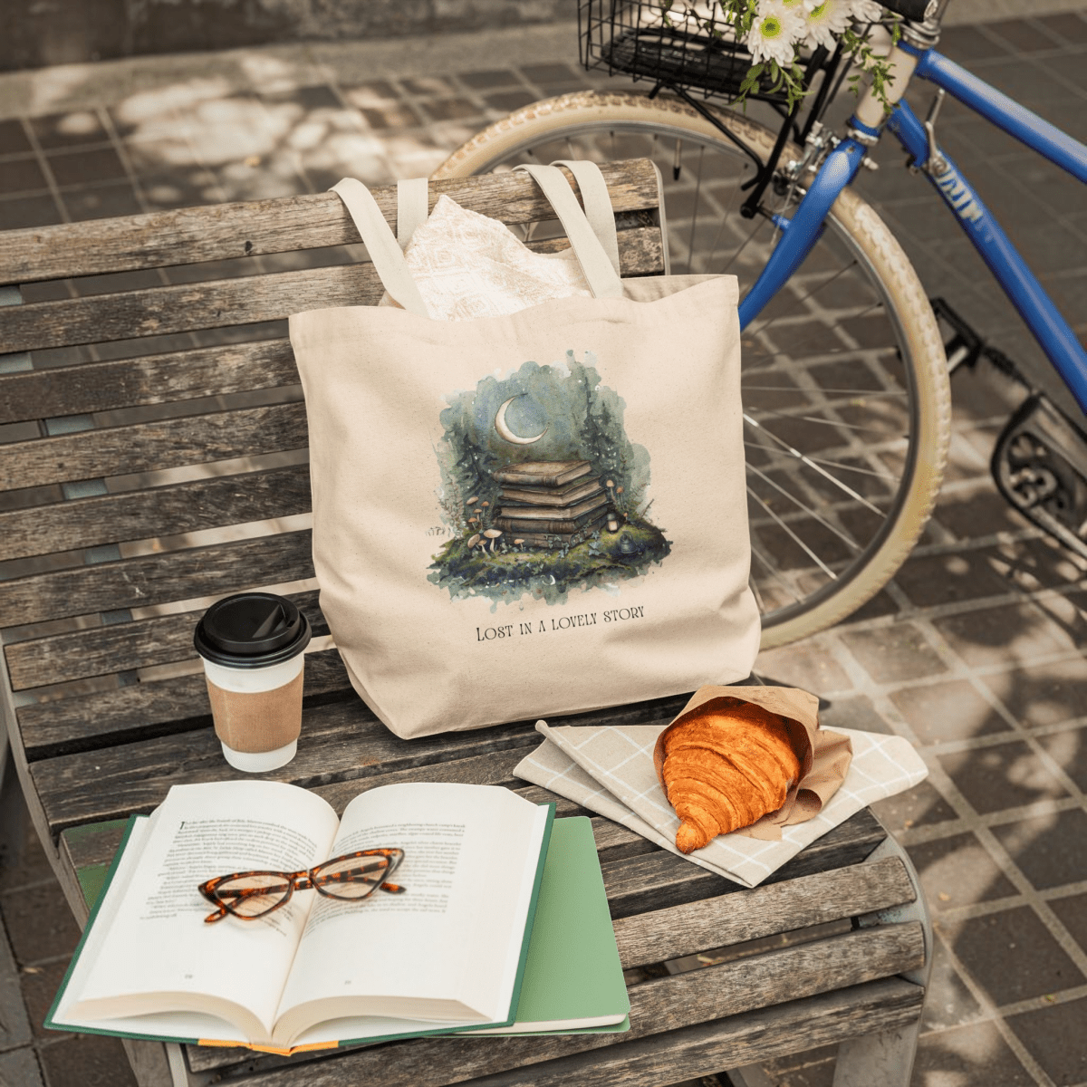 A Lost in a Lovely Story Canvas Tote Bag from Mirabilia Boutique rests on a bench alongside a coffee cup, croissant, open book with glasses, and a bicycle in the background. The canvas tote features the phrase "Lost in a lovely story," making it an ideal gift for any book lover.