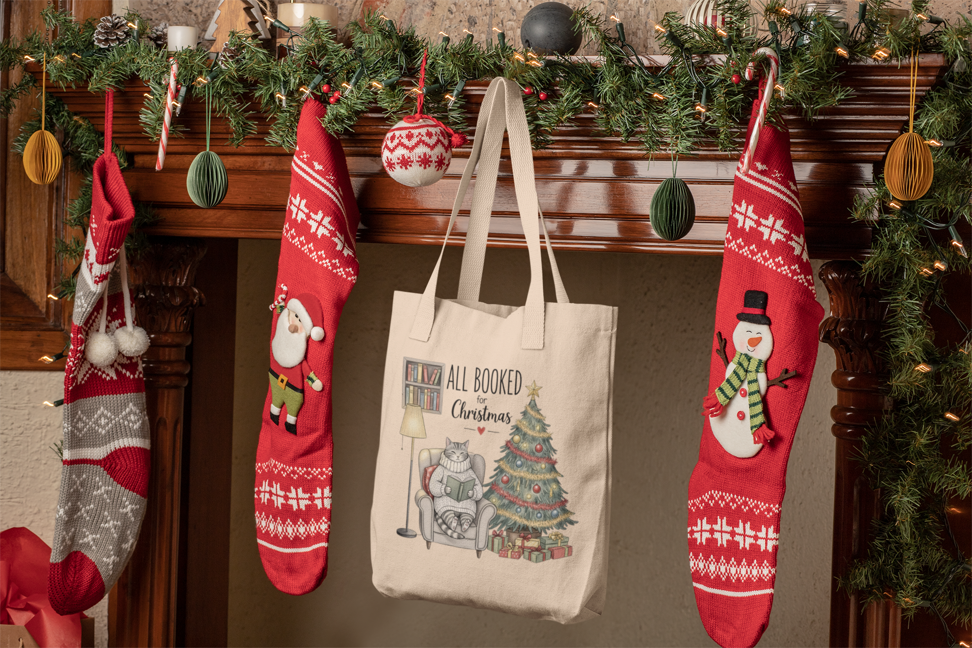 A festive fireplace mantel displays garland, ornaments, and red stockings. Centered is Mirabilia Boutique’s “All Booked for Christmas Cat Tote Bag”—a charming book lover’s gift featuring books, a tree, and a cute cat.