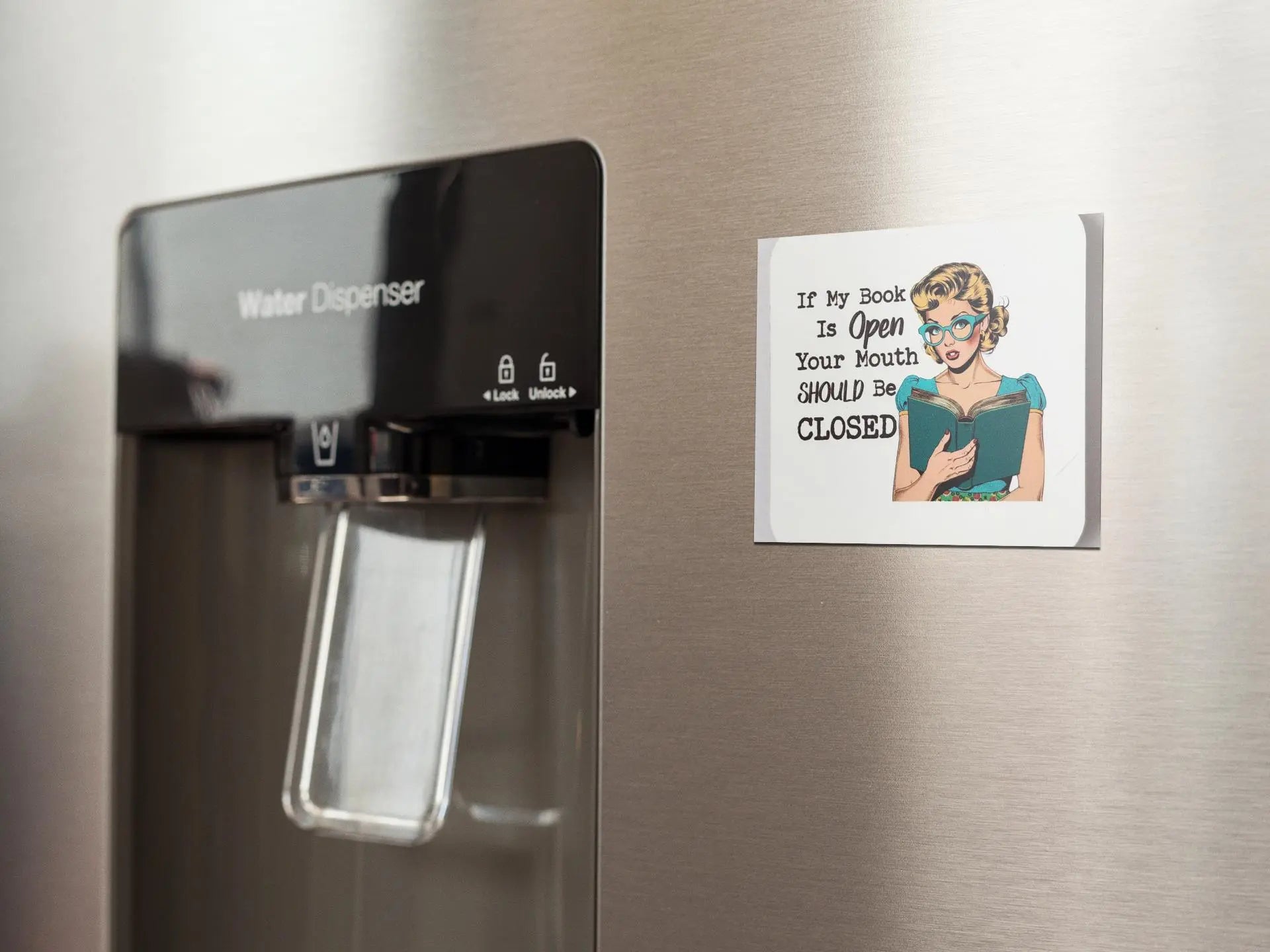 A refrigerator with a built-in water dispenser. To the right, a retro magnet from Mirabilia Boutique features an illustration of a woman in glasses holding an open book and the text: "If My Book is Open Your Mouth Should Be Closed." This makes for a perfect book lover gift, celebrating literary passion in style.