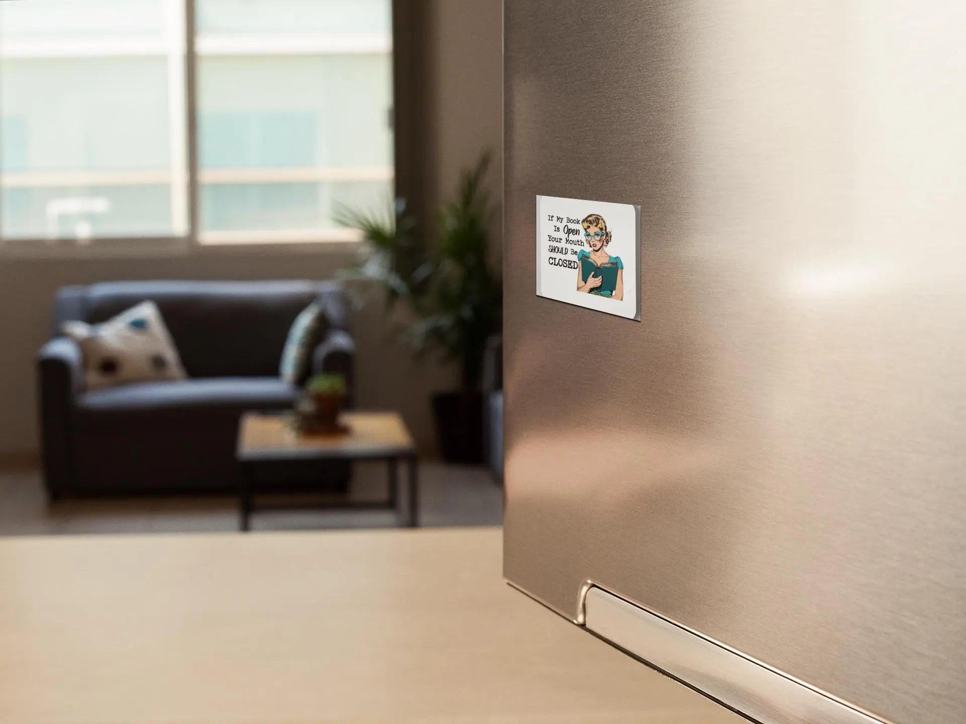 A kitchen scene with a refrigerator door in the foreground, displaying a retro magnet featuring a cartoon character and the text "If My Book is Open Your Mouth Should Be Closed" from Mirabilia Boutique. In the background, there's a cozy living area with a sofa, decorative pillows, and a coffee table graced by a plant that any book lover would delight in. Large windows let in natural light.