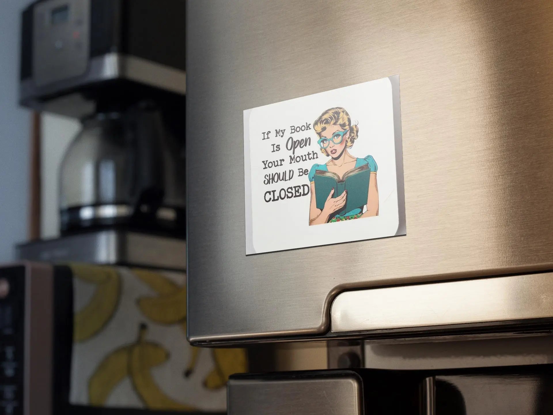 An "If My Book is Open Your Mouth Should Be Closed" magnet note from Mirabilia Boutique is attached to the refrigerator, featuring an illustration of a woman in glasses deeply engrossed in a book. The text whimsically warns, "If My Book Is Open Your Mouth SHOULD BE CLOSED." In the background, there is a coffee machine and wallpaper adorned with a banana pattern, making it an ideal gift for any book lover or literary passion enthusiast.
