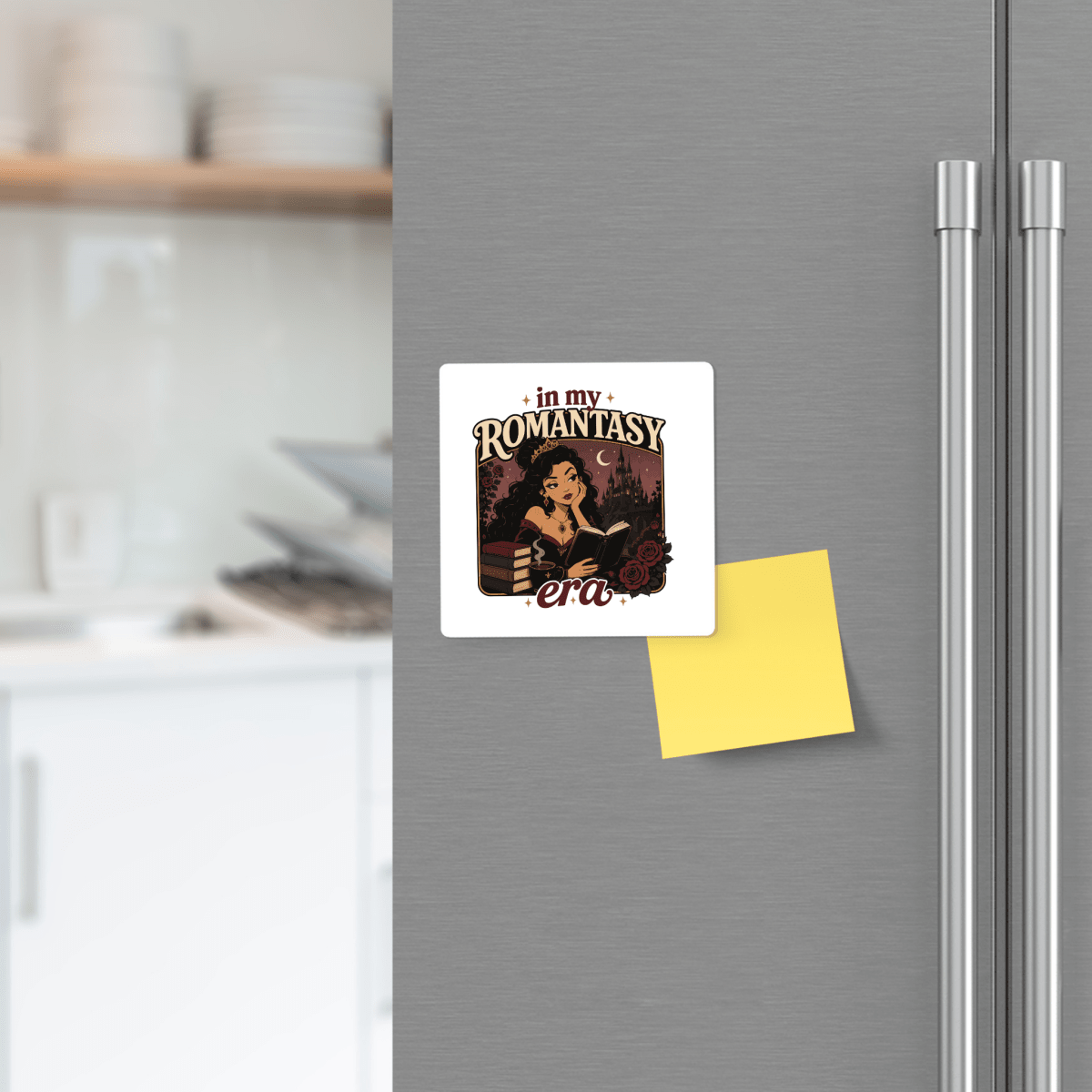 The "In My Romantasy Era Magnet" by Mirabilia Boutique—a square magnet featuring a woman reading and the phrase "in my ROMANTASY era"—is showcased on a gray refrigerator. Perched above a yellow sticky note, this delightful accessory adds a touch of bookish charm, with softly blurred kitchen shelves and dishes in the background, making it an ideal fantasy reader gift for any cozy home.
