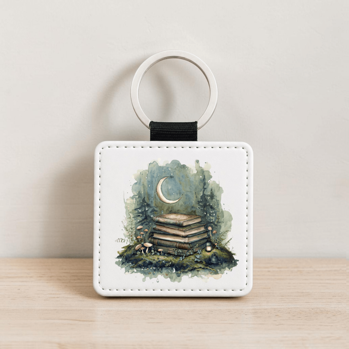 Introducing the Bookish Woodland Keychain by Mirabilia Boutique—a square keychain adorned with a whimsical illustration of stacked books, mushrooms, and a glowing crescent moon nestled among woodland trees. Set on a light wood surface against a simple background, this enchanting cottagecore accessory is perfect for book lovers and nature enthusiasts alike.