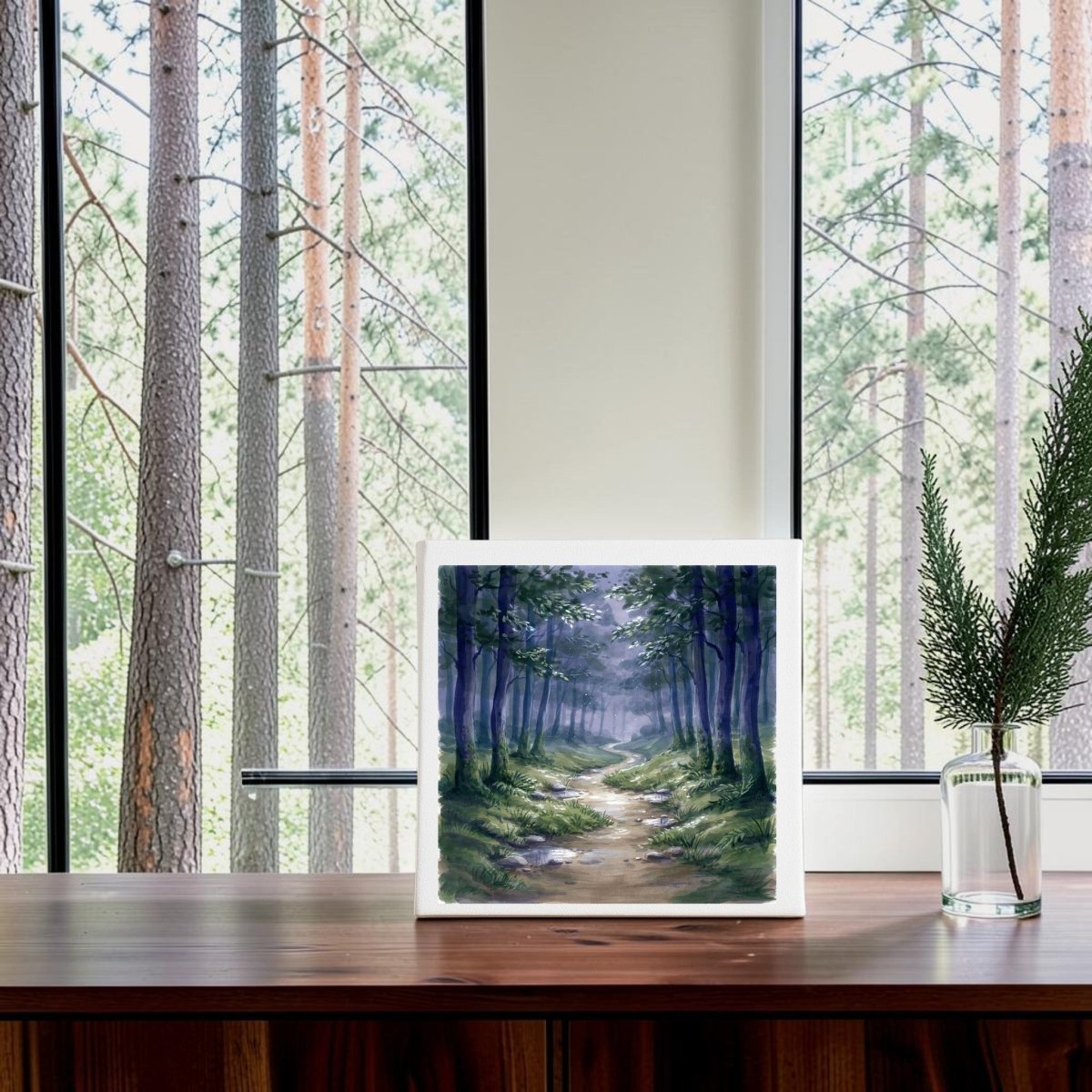 The Borrow a Little Moonlight Forest Canvas Wall Art by Mirabilia Boutique rests on a wooden table near a window, with tall pine trees visible outside. A glass vase filled with green foliage sits on the right side of the table, enhancing the serene atmosphere created by this tranquil decor piece.