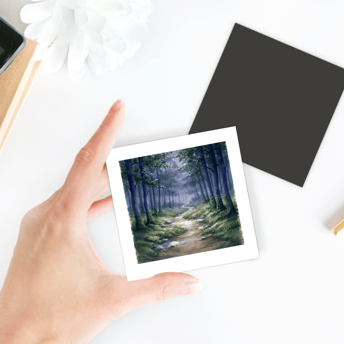 A hand holds the Borrow a Little Moonlight Forest Magnet by Mirabilia Boutique, featuring a square photo of a serene forest path surrounded by trees. The miniature artwork, designed as a decorative magnet, is displayed on a white surface alongside a black square and a white flower.