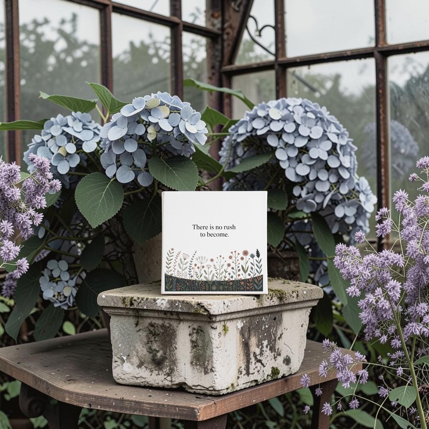 The "There Is No Rush to Become" Canvas Art by Mirabilia Boutique rests on a weathered stone planter with hydrangeas and lavender—an inspiring accent for motivational home decor or a thoughtful gift.