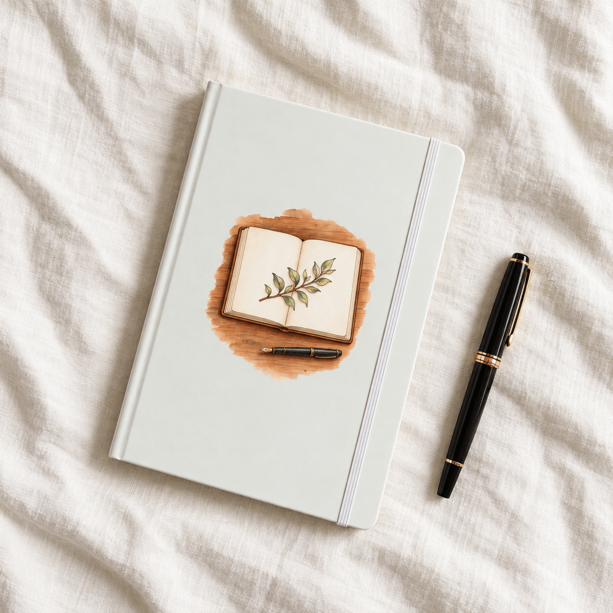 A white A5 Living Page Botanical A5 Journal by Mirabilia Boutique featuring a watercolor cover illustration of an open book with a leafy branch, lying next to a black fountain pen on a white linen surface.