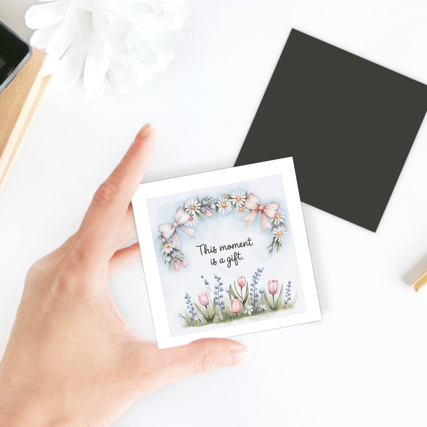 A hand holds the "This Moment Is a Gift Magnet – Floral Spring Quote" by Mirabilia Boutique, featuring floral art and uplifting words—a perfect inspirational gift for daily mindfulness. Nearby are a black envelope, white flower, and notebook.