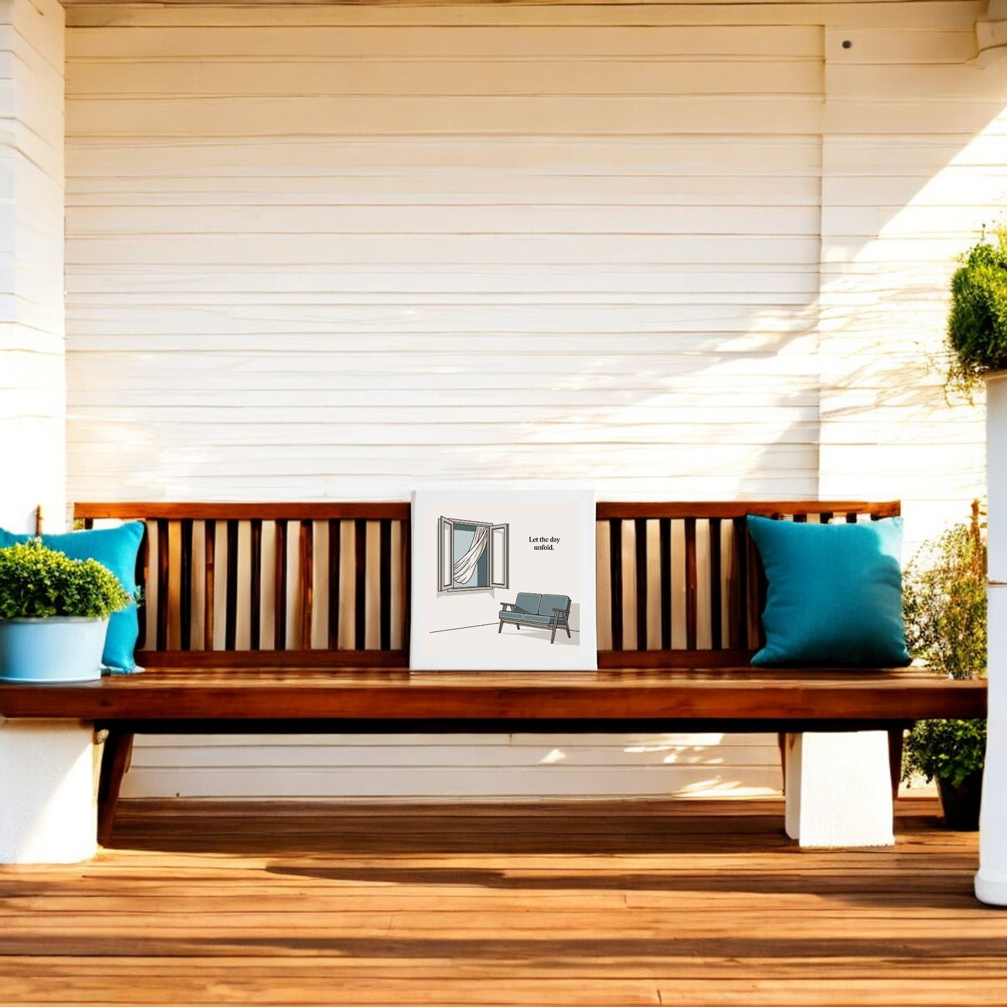 A Mirabilia Boutique "Let the Day Unfold Canvas – Serene Slow Morning Art" hangs above a wooden bench with blue cushions on a porch, while potted plants enhance the relaxing, slow-living atmosphere.