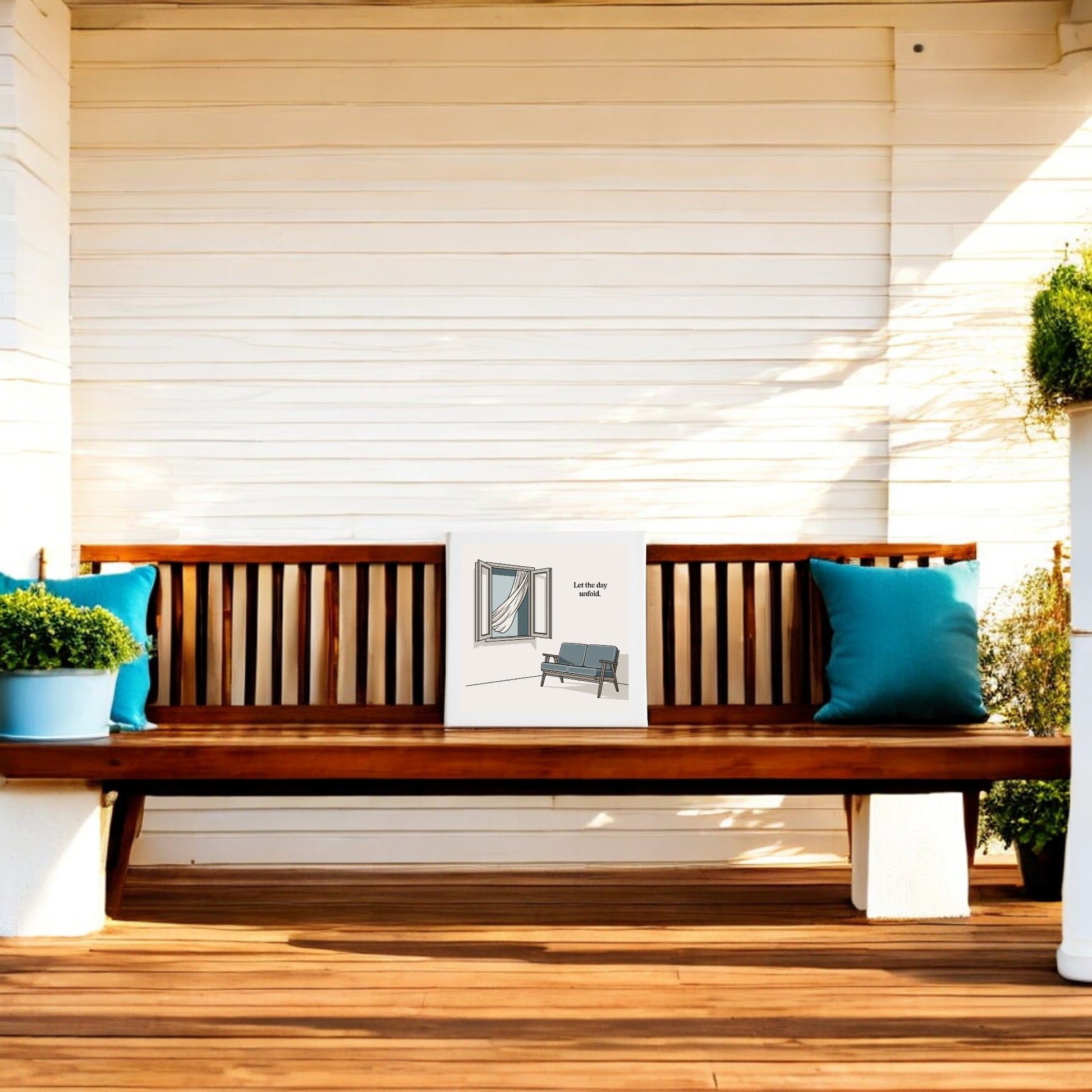 A Mirabilia Boutique "Let the Day Unfold Canvas – Serene Slow Morning Art" hangs above a wooden bench with blue cushions on a porch, while potted plants enhance the relaxing, slow-living atmosphere.