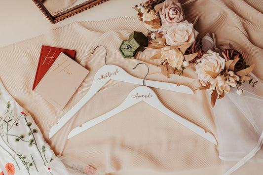 Flat lay of Mirabilia Boutique’s Personalized Wedding Hangers | Custom Name Hanger Set, labeled "Ashley" and "Anahi," styled with florals, a veil, embroidered fabric, a ring box, and vow books titled "His" and "Hers.